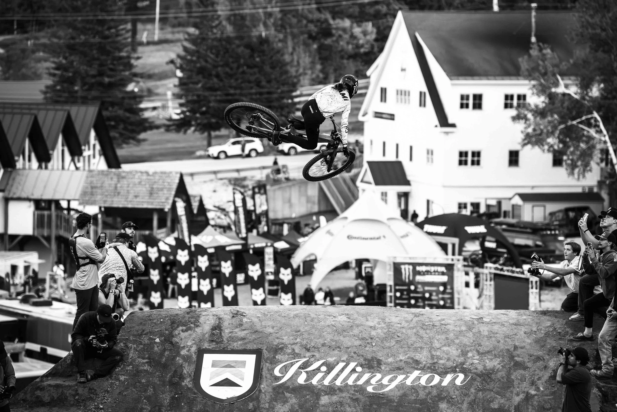 A mountain biker performing a backflip in mid-air during a competition, with spectators and photographers capturing the stunt, in Killington with a backdrop of tents, buildings, and trees.