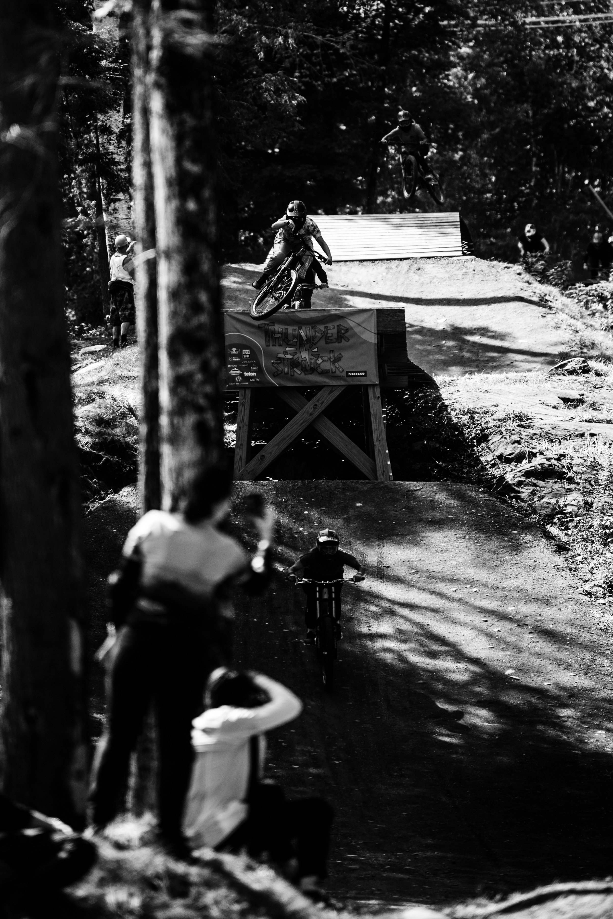 Mountain bikers performing jumps on a rugged trail in a forest during a downhill biking event, with spectators watching from the side.