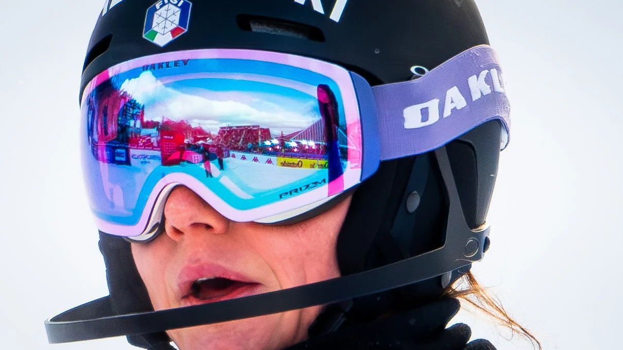 Close-up of a person wearing a black ski helmet and reflective ski goggles, with snow in the background.