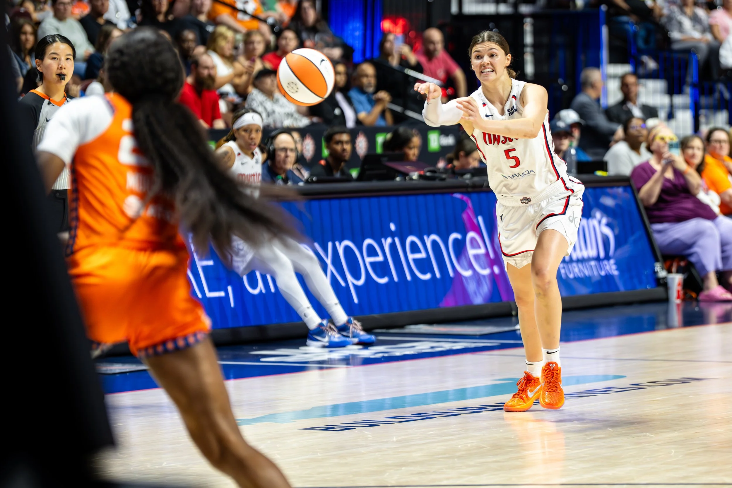 A basketball player in a white jersey with red and blue details prepares to pass the ball during a game. She is wearing orange shoes and is positioned near the sideline. An opponent in an orange uniform is seen in the foreground, blurred, while fans 