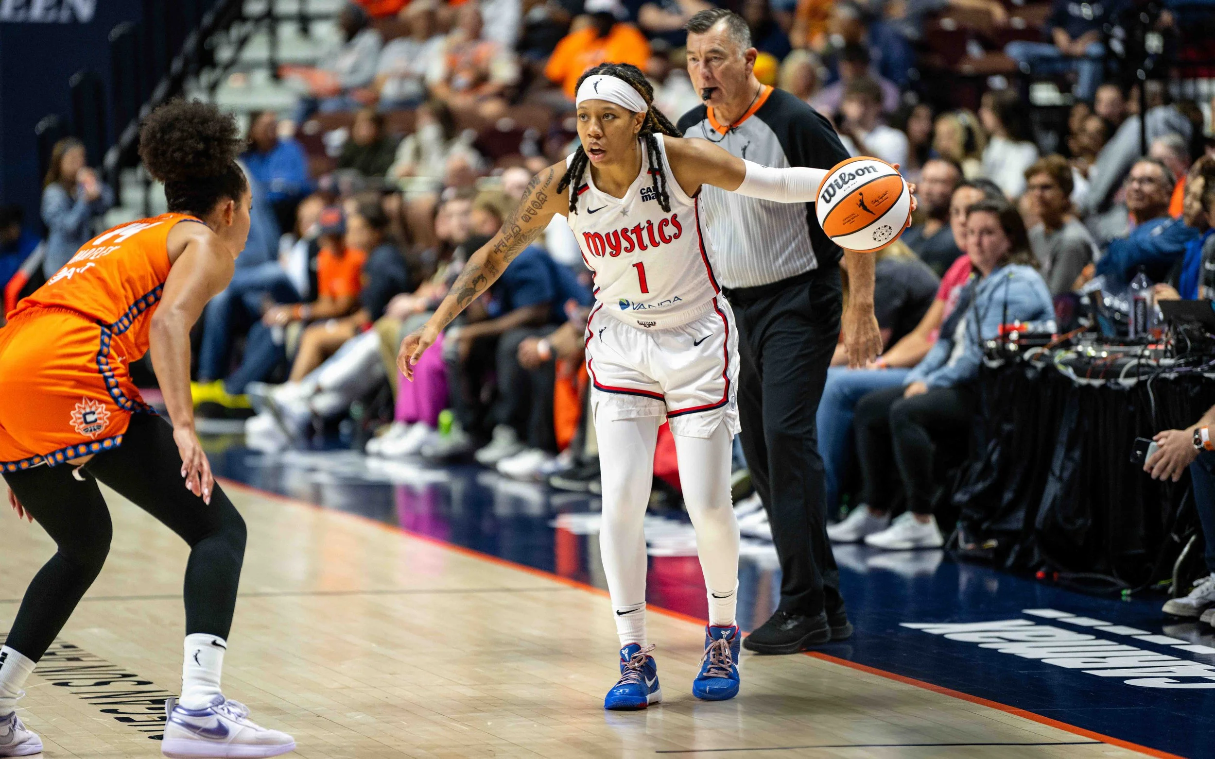 A basketball player in a white jersey labeled 'Mystics' with the number 1, dribbling the ball as a defender in an orange jersey attempts to guard her. The game is taking place indoors with spectators in the background.