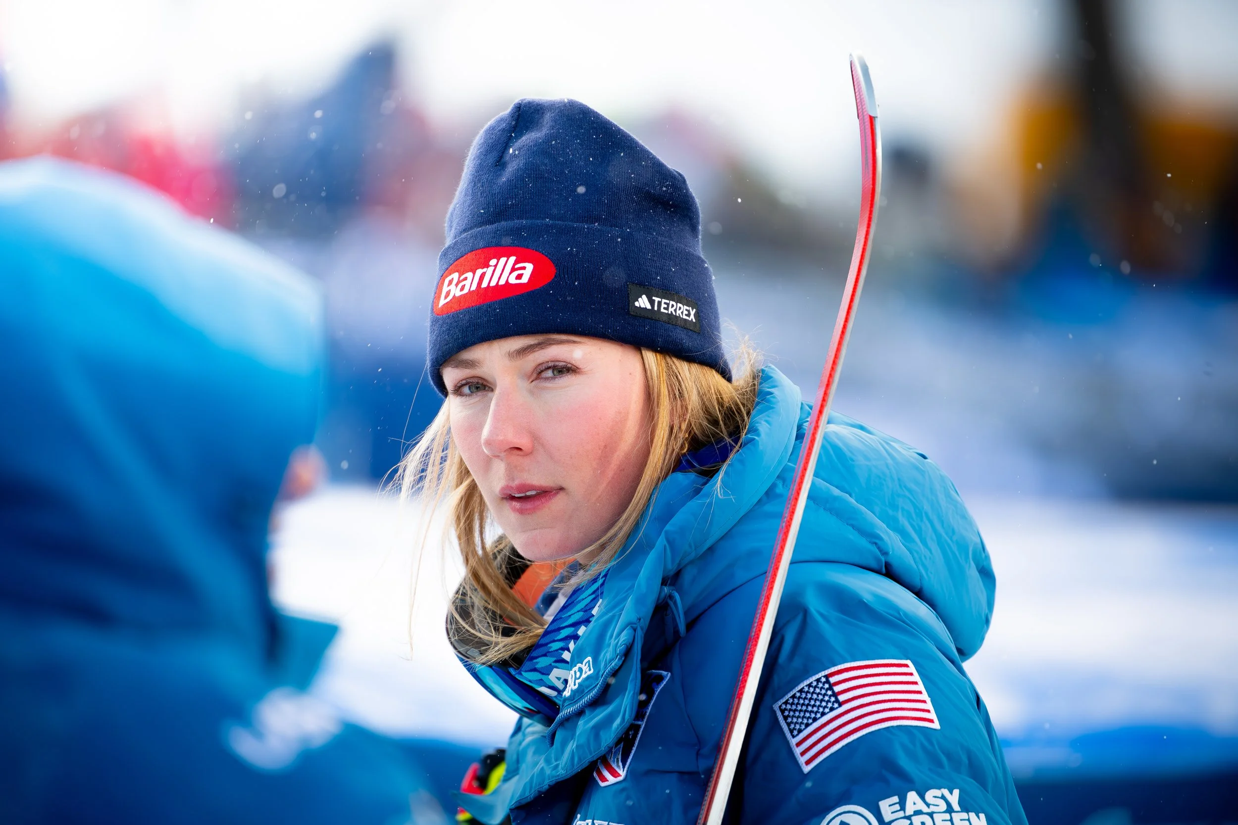 A female athlete in winter gear, wearing a blue jacket with an American flag patch, a navy beanie with logos, and holding a ski, outdoors in a snow setting.