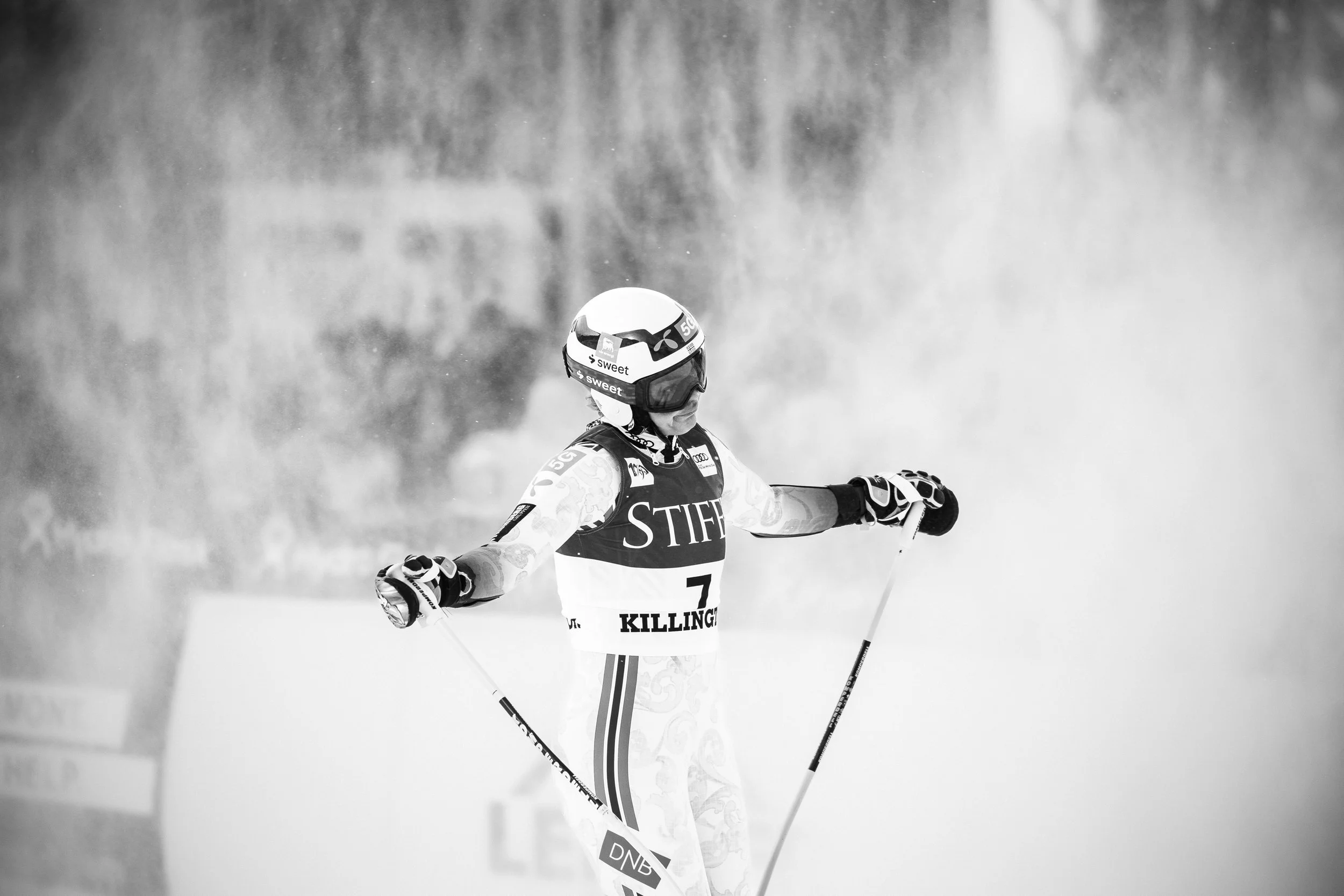A skier in racing gear and helmet on snow with arms outstretched, creating snow spray during a ski race.