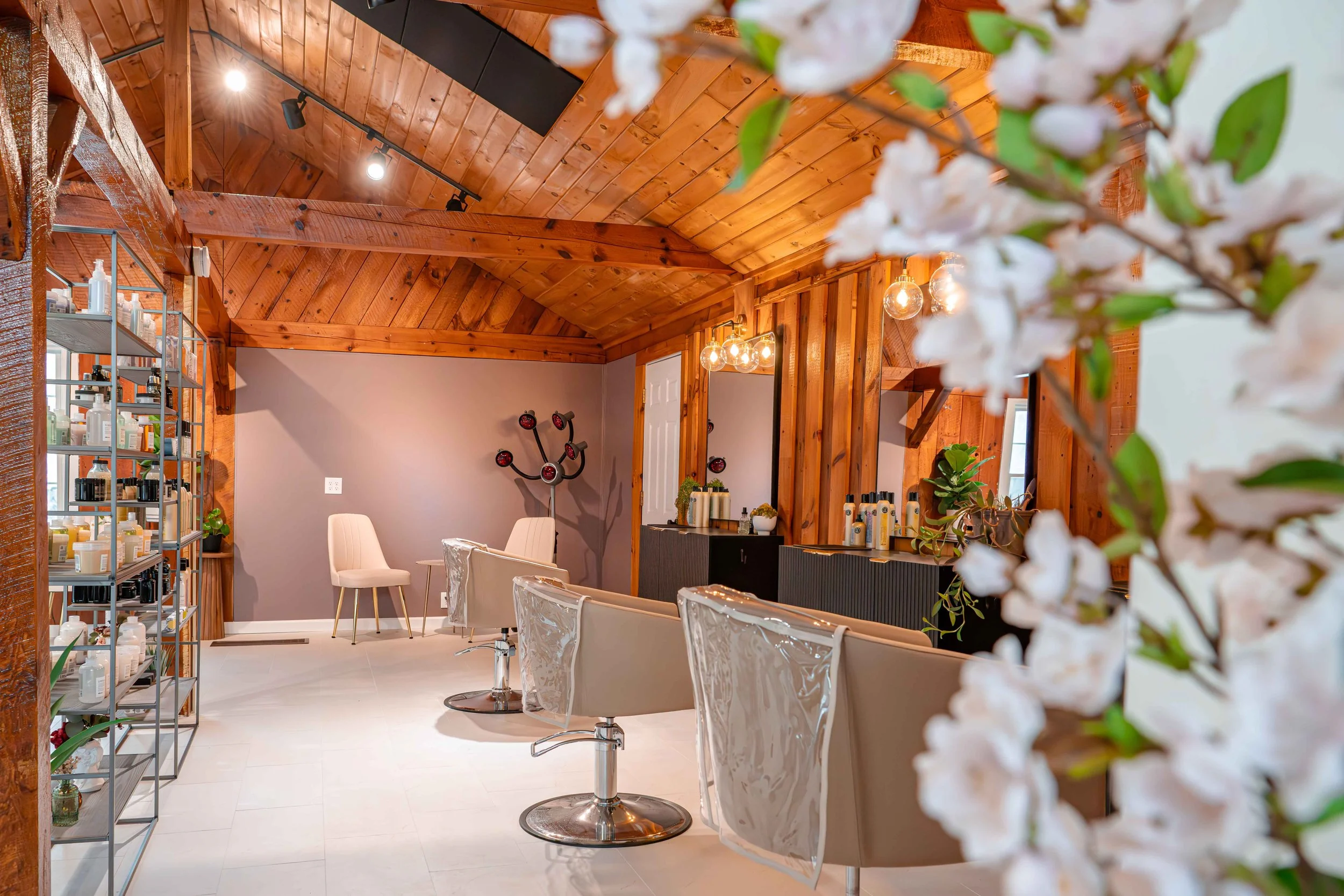 A modern salon with wooden vaulted ceiling, styling chairs, a mirror, and shelving with hair products, decorated with a branch of white flowers in the foreground.