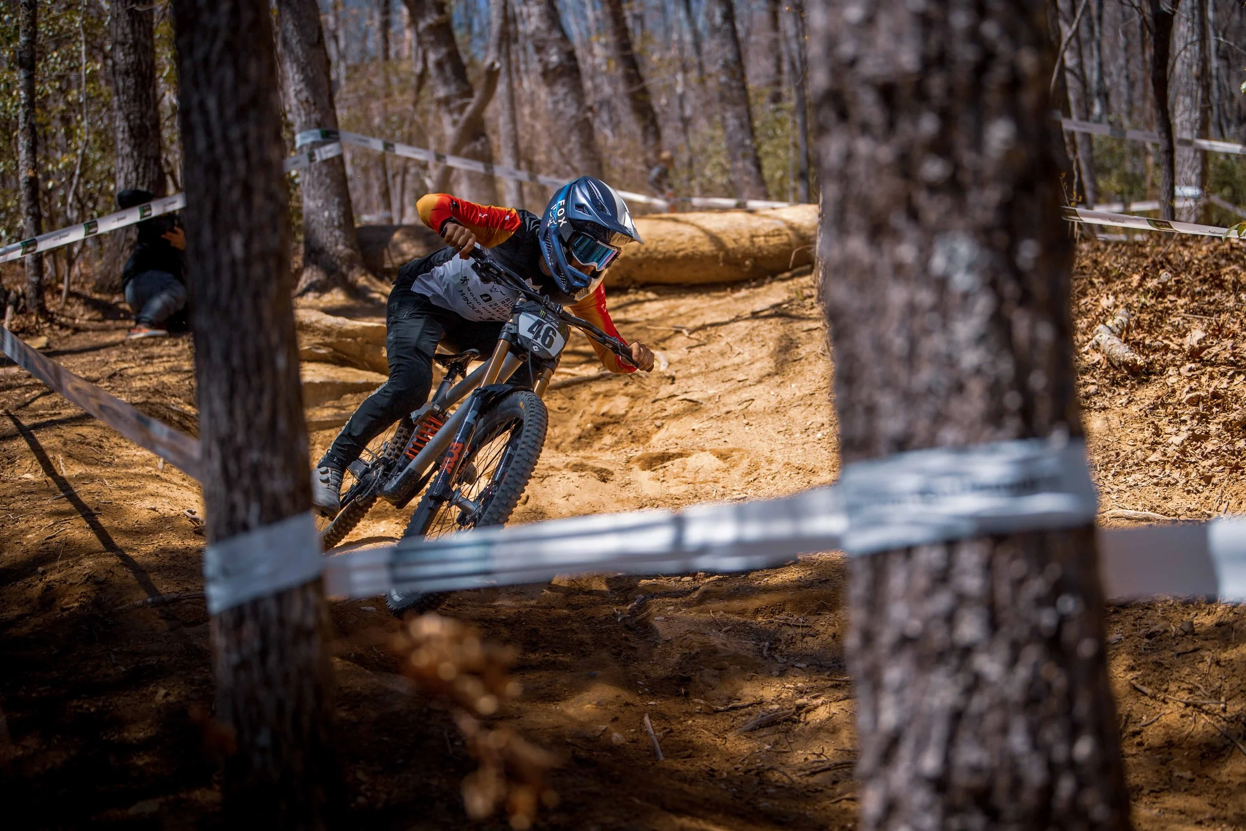 A mountain biker in mid-air riding along a dirt trail through a wooded area, wearing a helmet, goggles, and riding gear, with trees and safety tape lining the course.