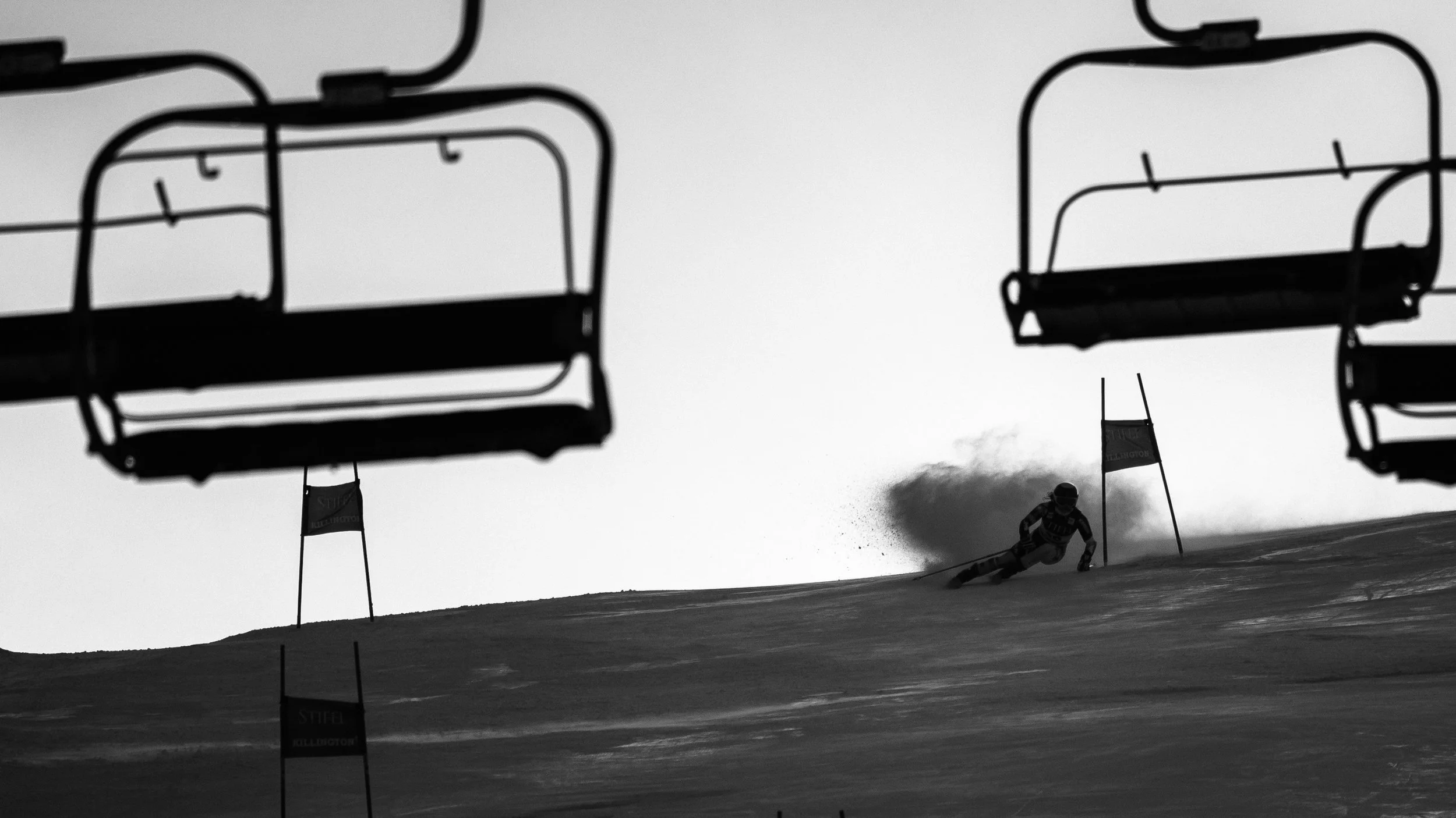 A skier racing downhill past slalom gates on a mountain slope, with empty ski lift chairs in the foreground and snow spray behind the skier.