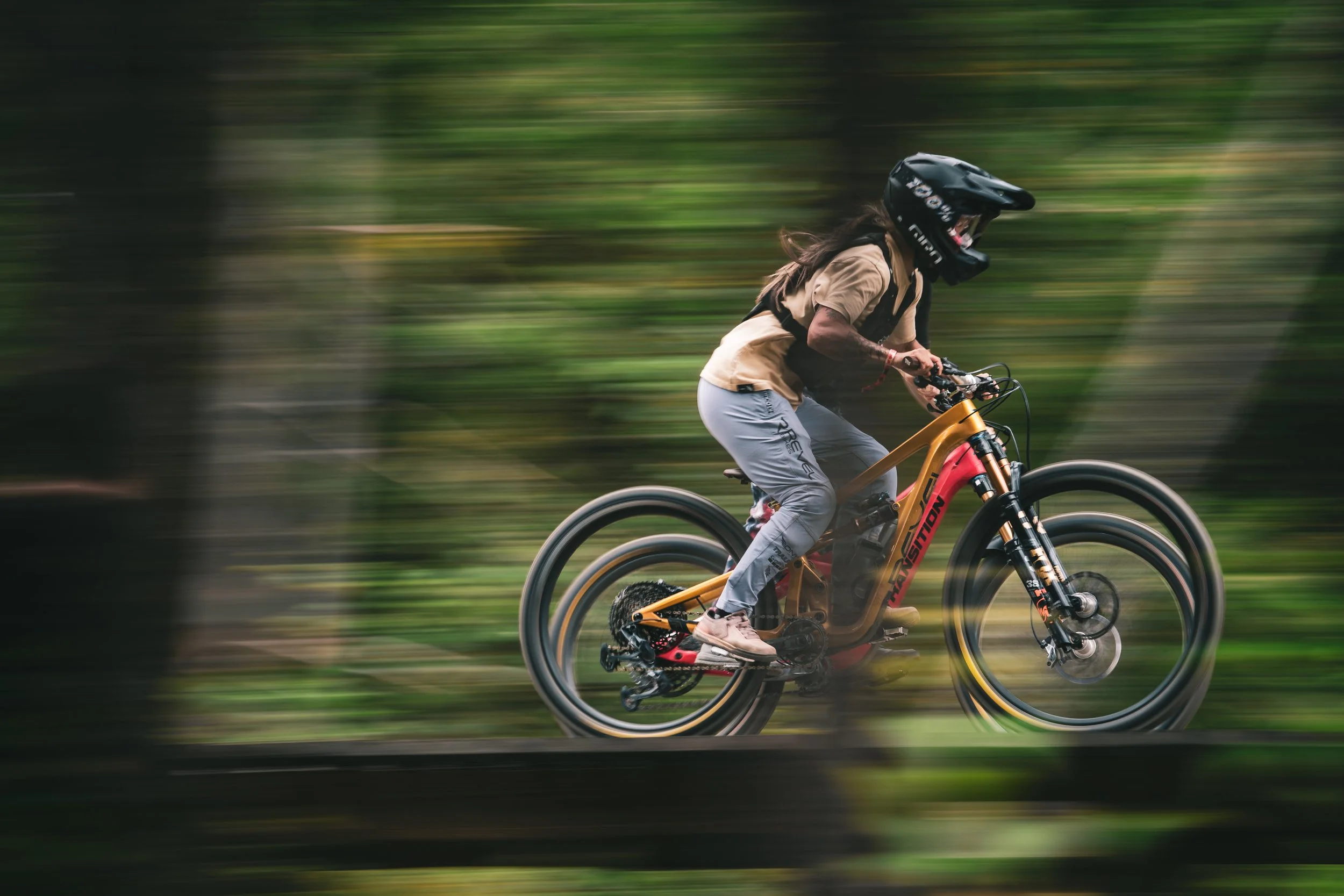 A person riding a mountain bike through a forest, wearing a black helmet, beige shirt, grey pants, and sneakers, with a motion blur background indicating fast movement.