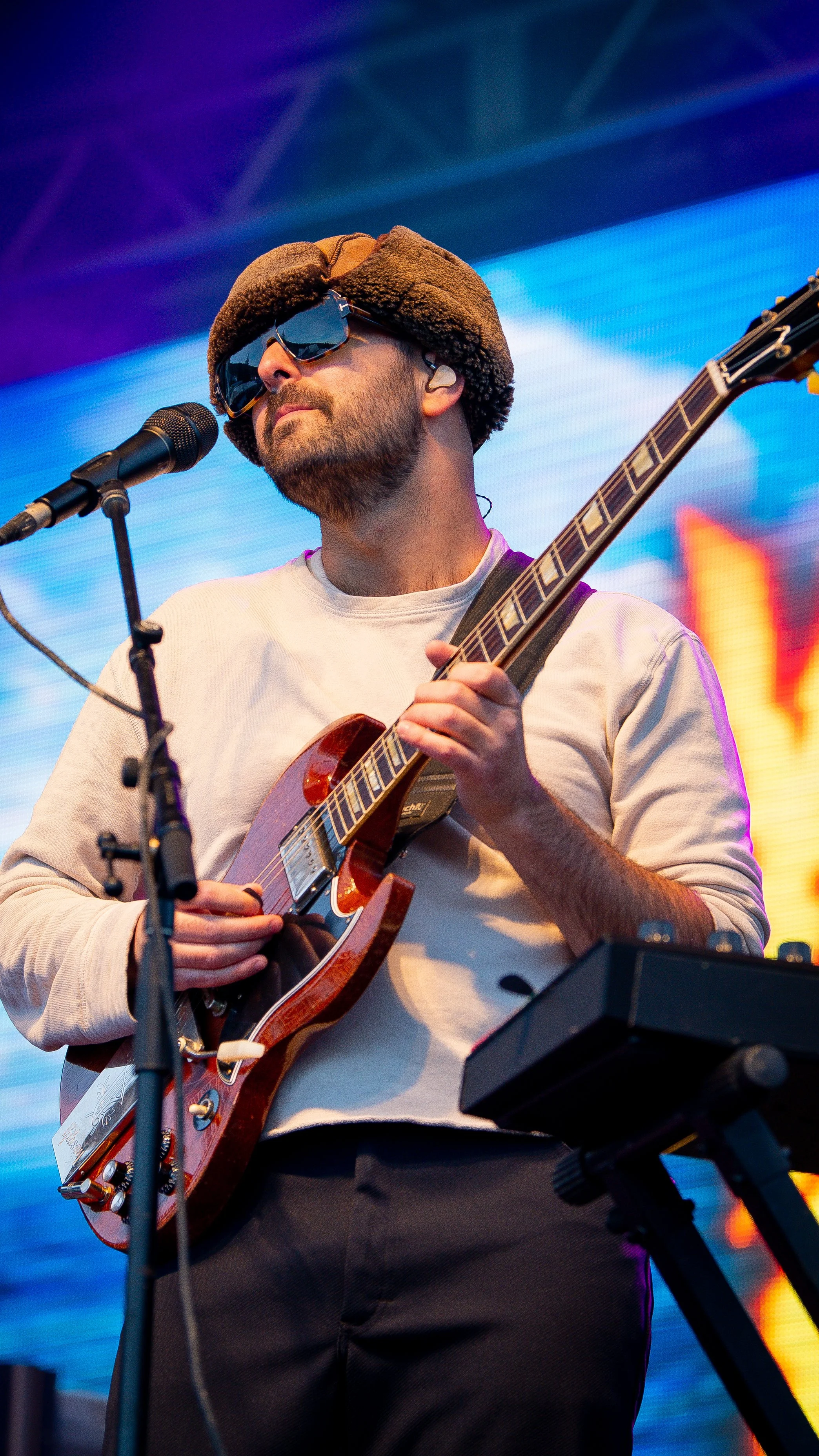A man wearing sunglasses, a furry hat, and a white long-sleeve shirt is playing an electric guitar and singing into a microphone on stage with a colorful background.