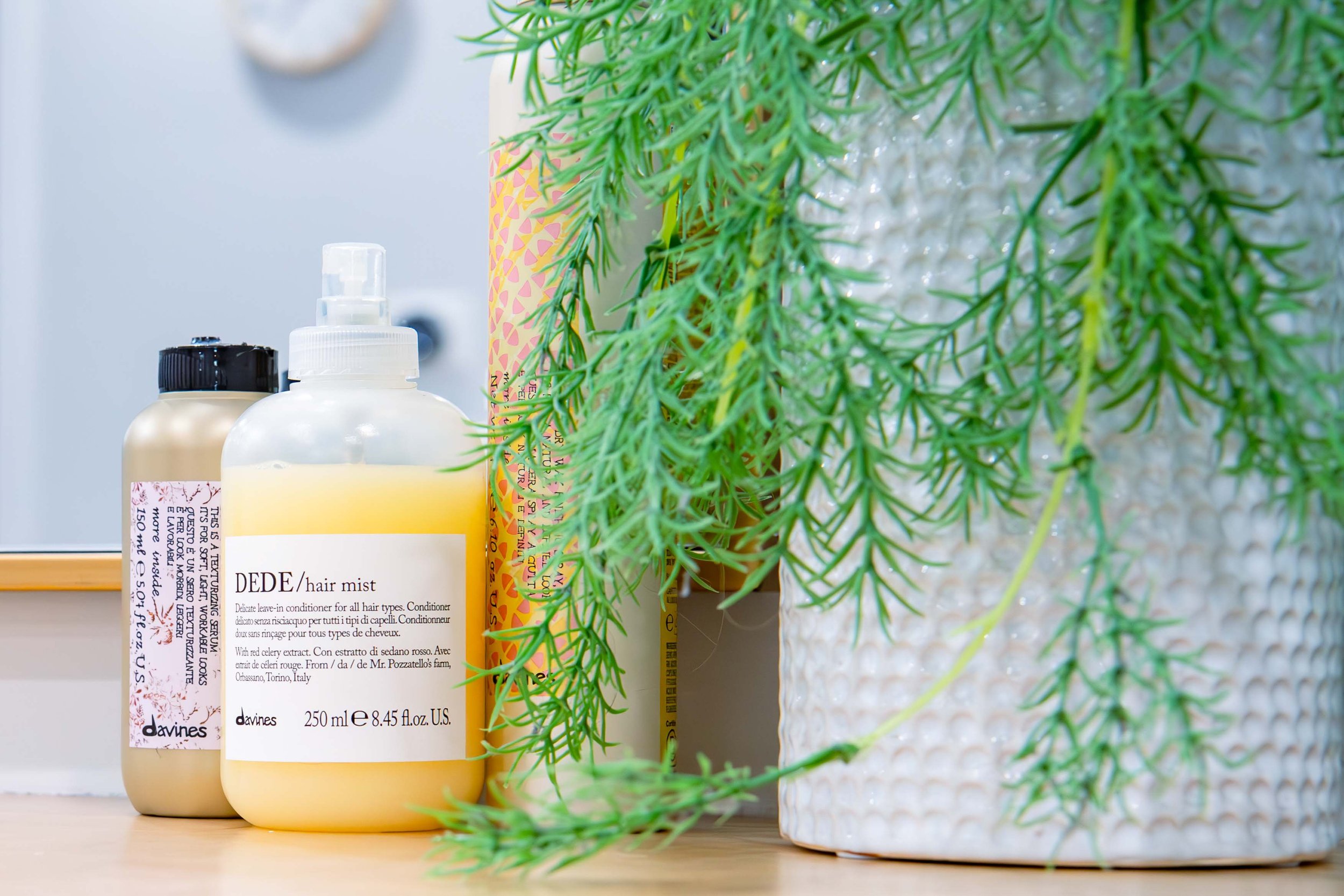 Hair care products, including a bottle of DeDE hair mist, placed beside a potted plant with green leaves on a wooden surface.
