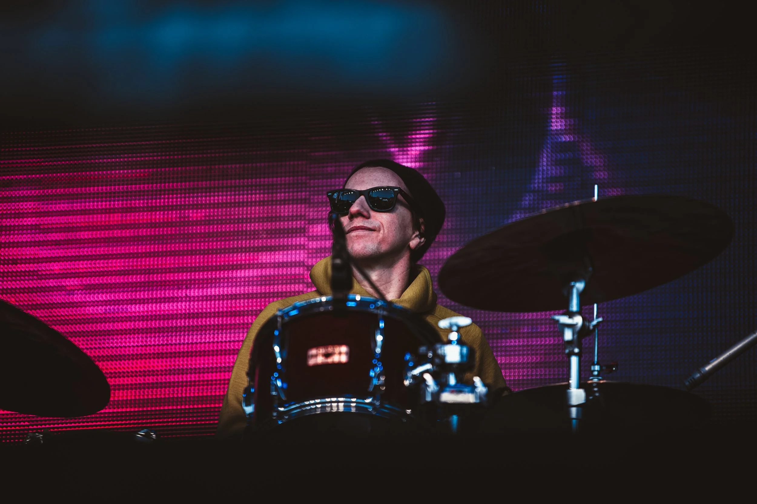 A man wearing sunglasses and a yellow hoodie playing drums on stage with a colorful LED background.