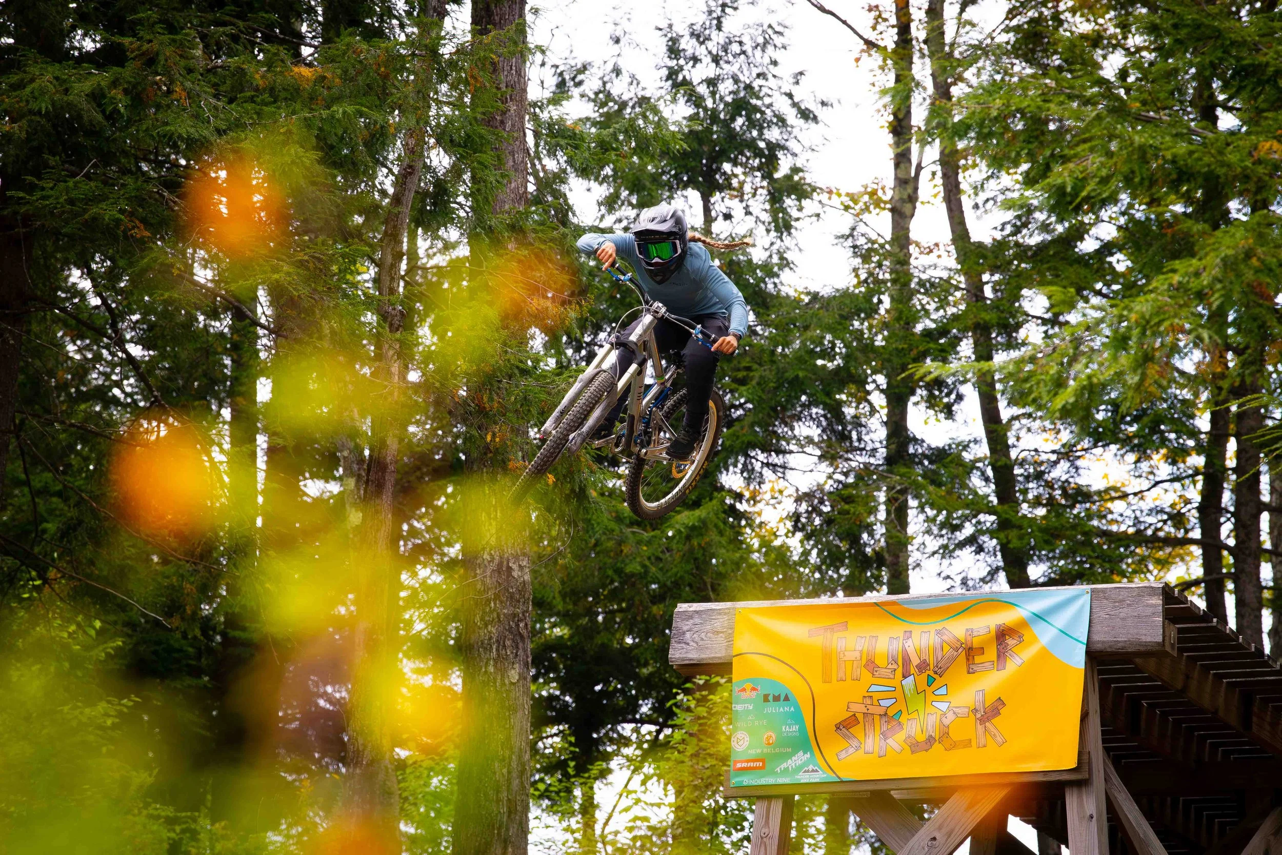A mountain biker in a helmet and protective gear jumps off a wooden ramp labeled "Thunder Struck" in a forested area.