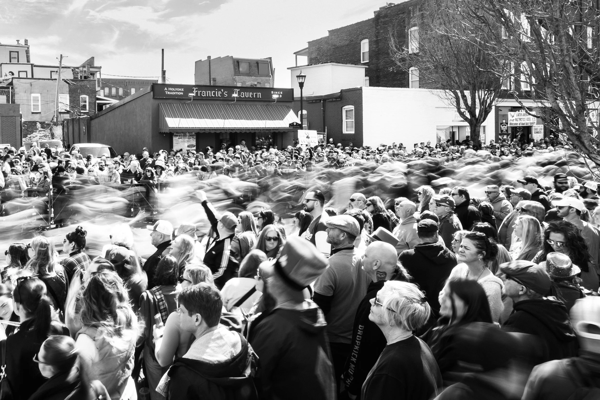 Black and white photo of a large crowd gathered outdoors in front of a tavern, with some motion blur indicating movement, and buildings in the background.