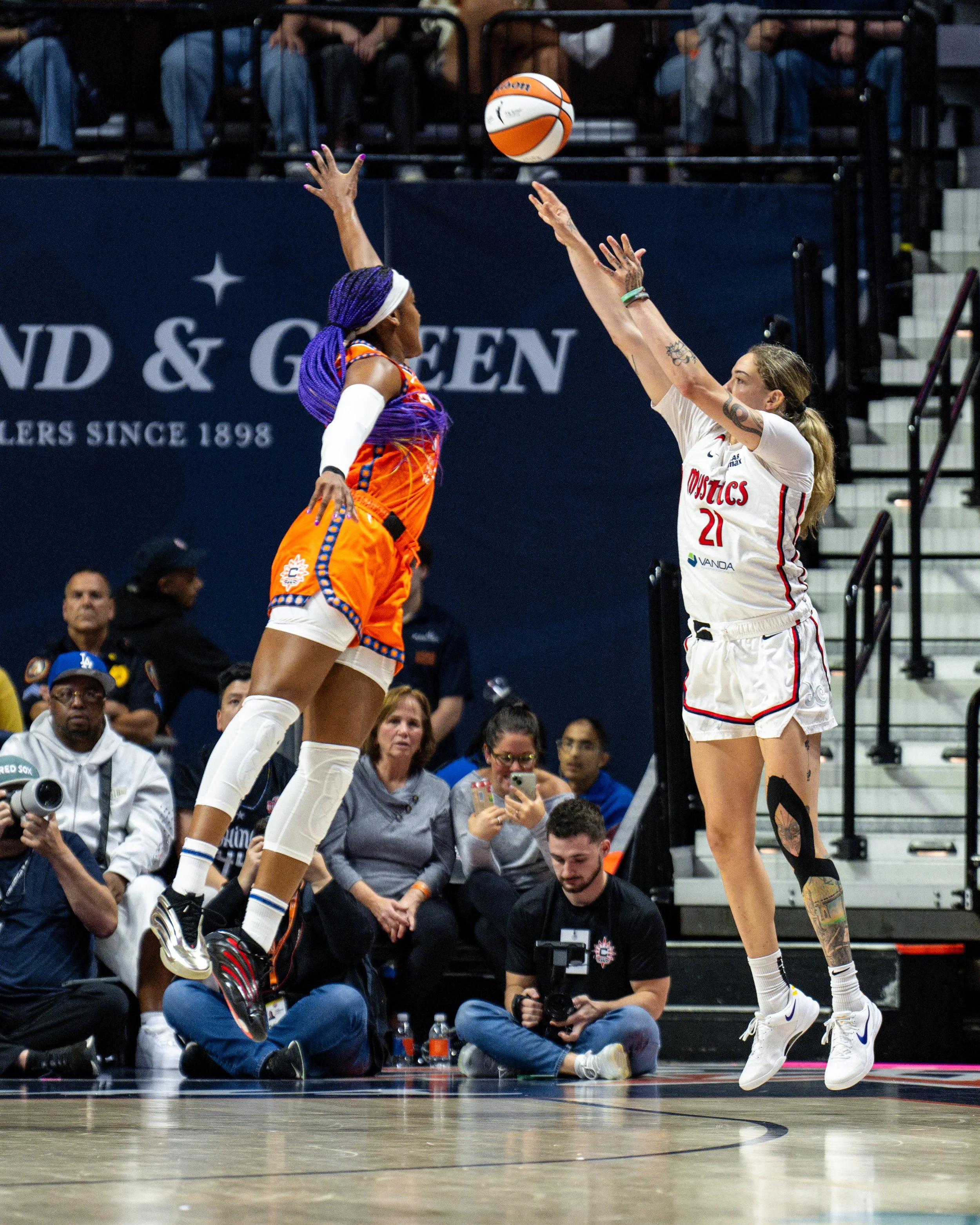 Two female basketball players in mid-air during a game, one in an orange uniform and the other in a white uniform, reaching for the ball. Spectators and photographers are visible in the background.
