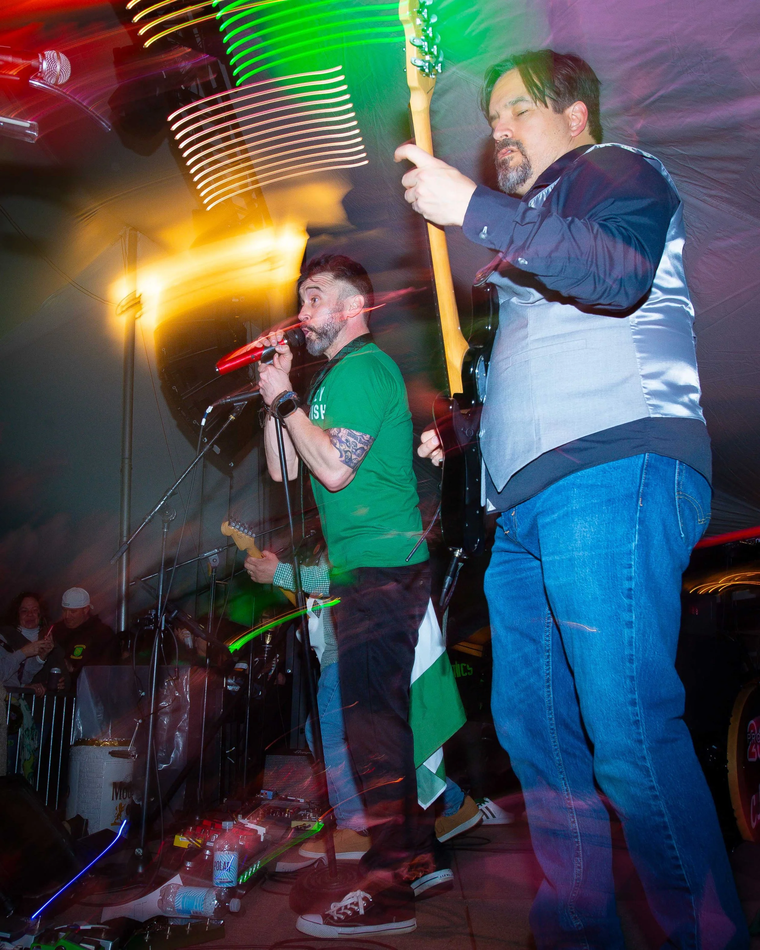 Musicians performing on stage with colorful lights, one singing into a red microphone and the other playing an electric guitar, audience watching in the background.