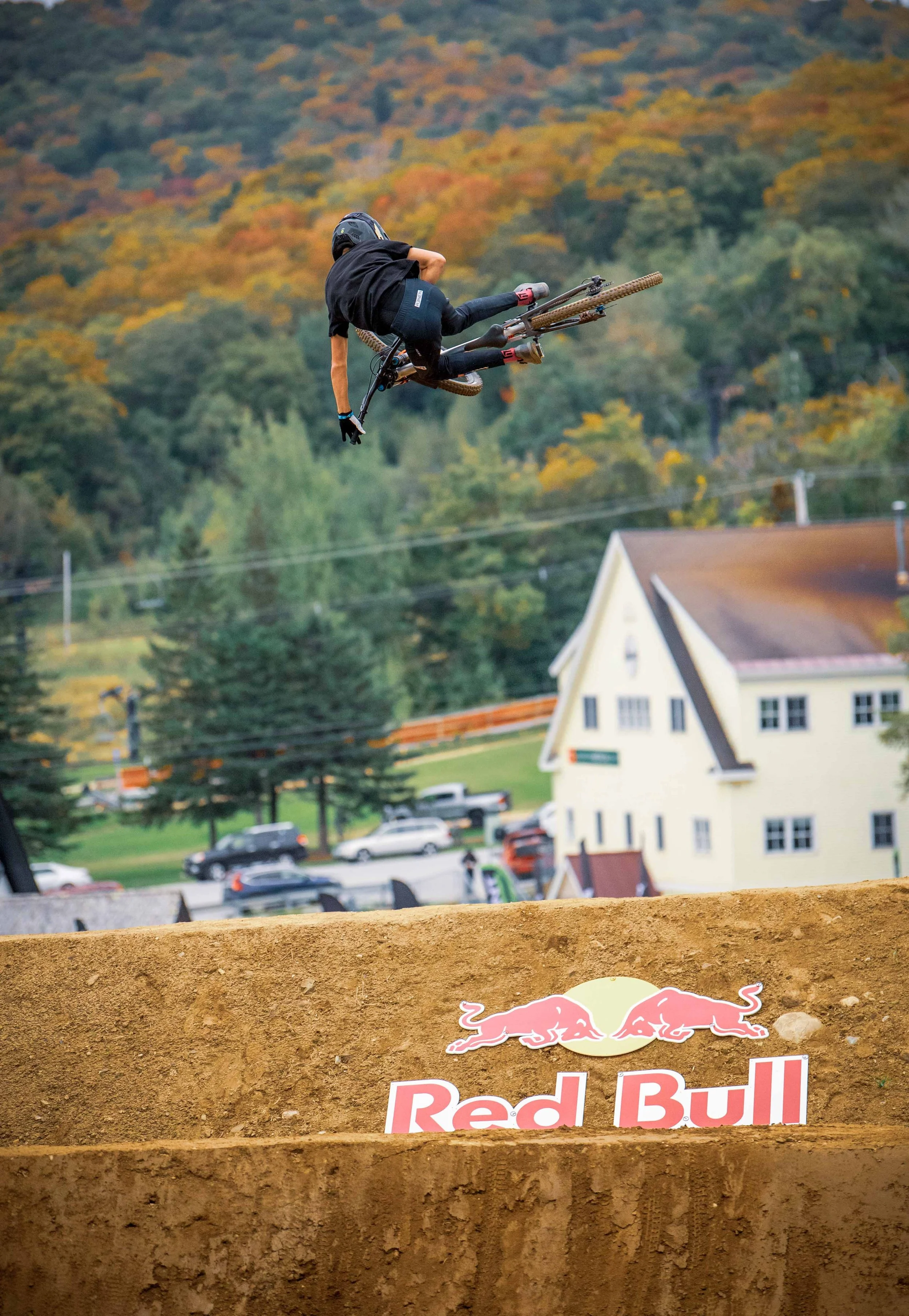A mountain biker in black riding gear performs a jump at a dirt track with a Red Bull logo, with a forested hillside and houses in the background.