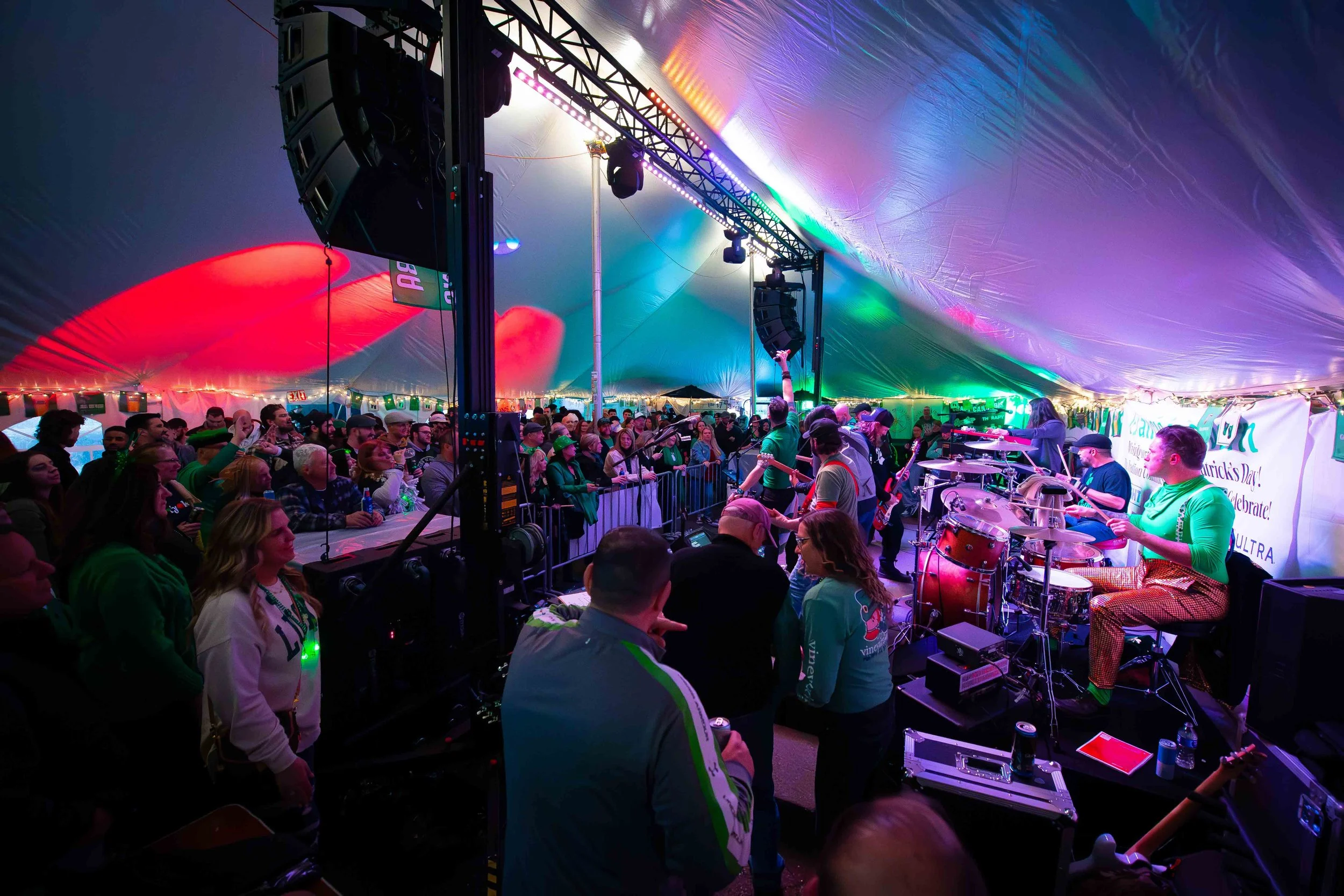 A lively crowd attending a concert inside a large tent decorated with colorful lights. On stage, a band performs with various instruments, including drums and guitars, while the audience enjoys the event.