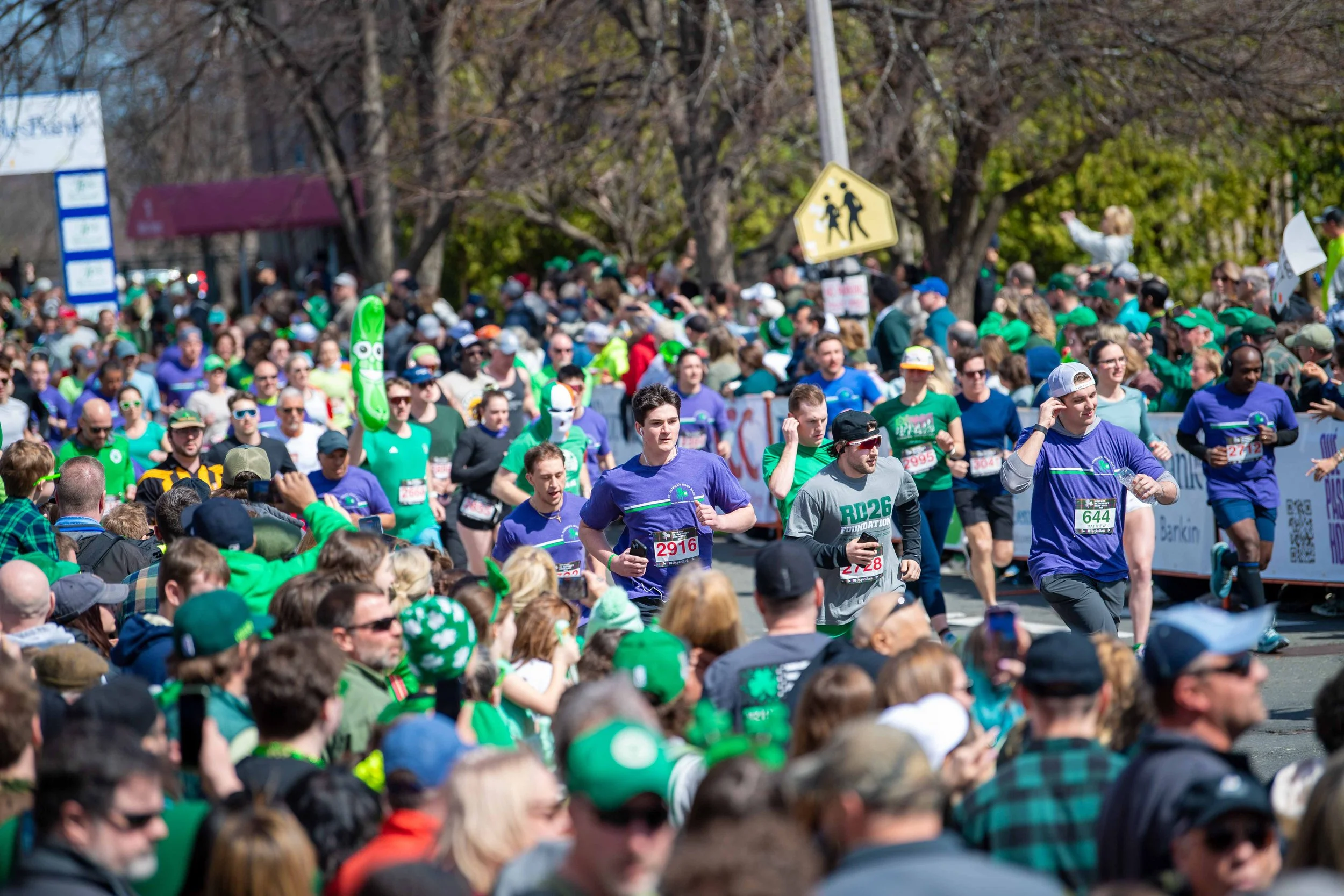 Crowd of people participating in a marathon, wearing colorful athletic clothing and race bibs, with some spectators watching and taking photos, set outdoors on a sunny day with trees and street signs in the background.