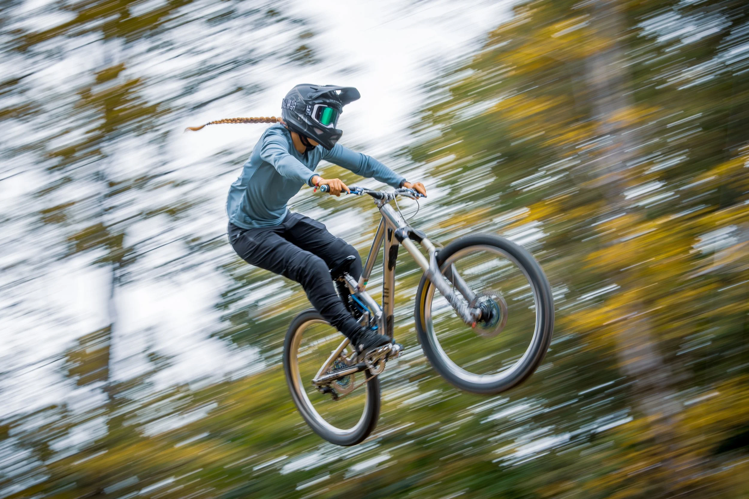 A person wearing a helmet and protective gear riding a mountain bike through a forest trail, captured mid-air in a jump with blurred background.