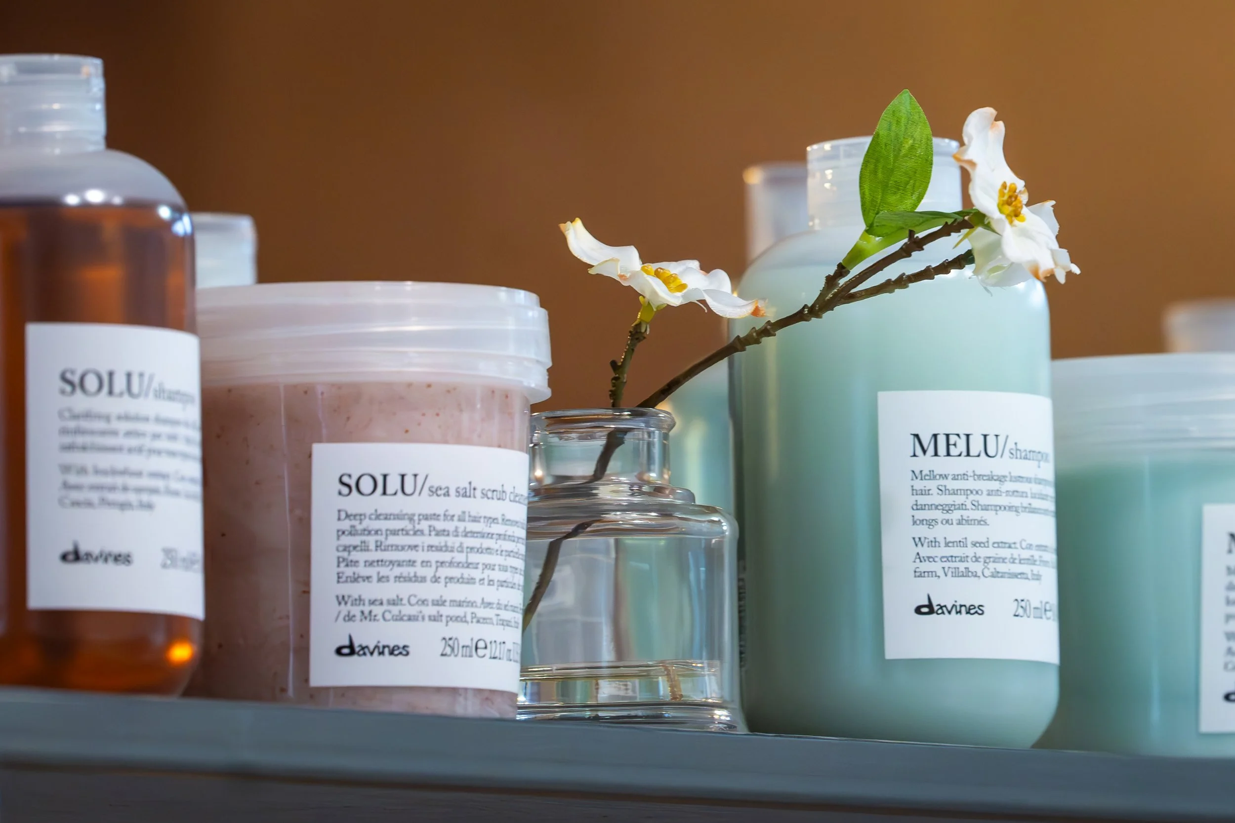 Collection of hair care products including shampoo, scrub, and spray bottles on a shelf with a flower branch.