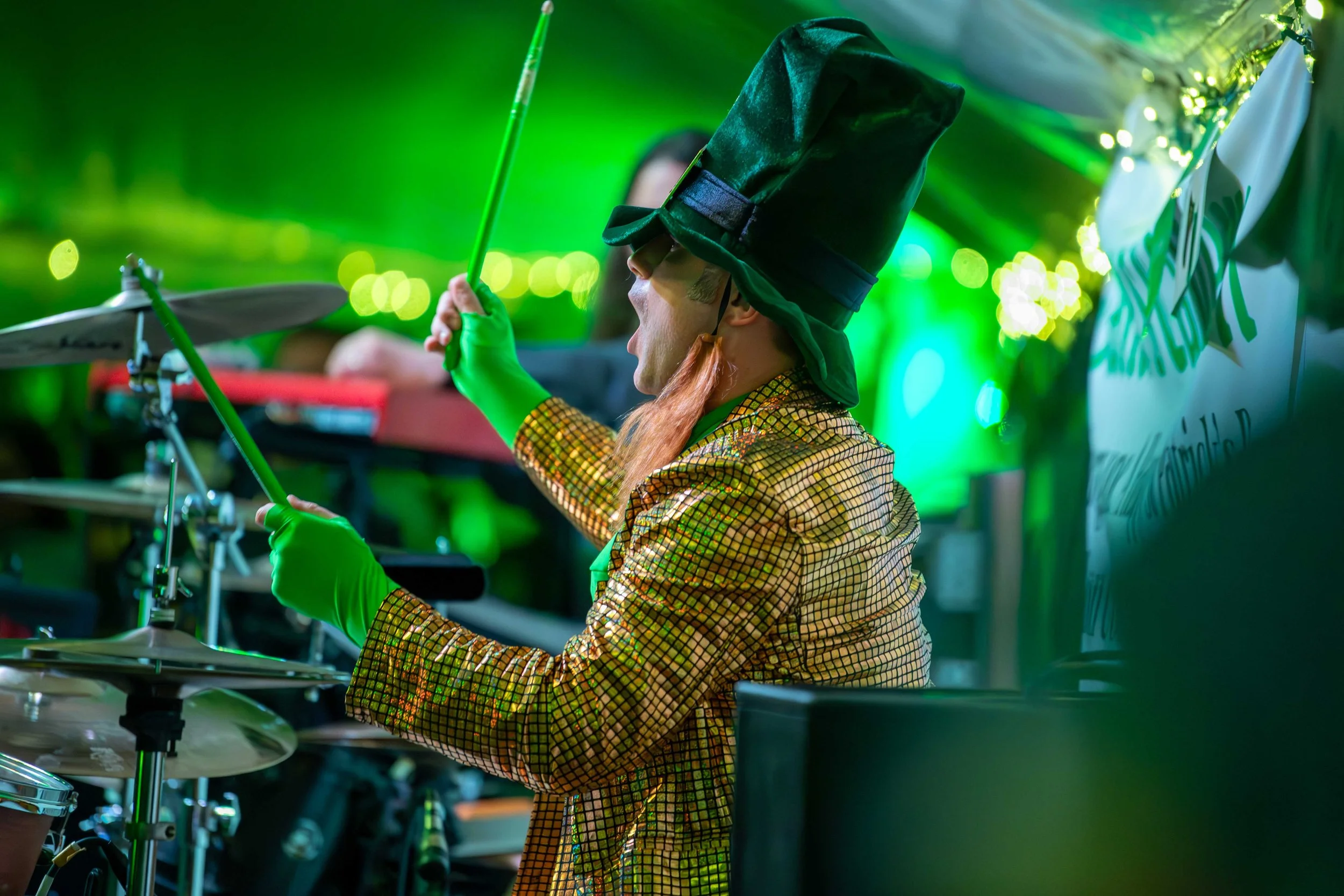 A person dressed as a wizard with a tall black hat, green gloves, and a shiny gold checkered jacket is playing the drums at a festive event, illuminated by green lighting.