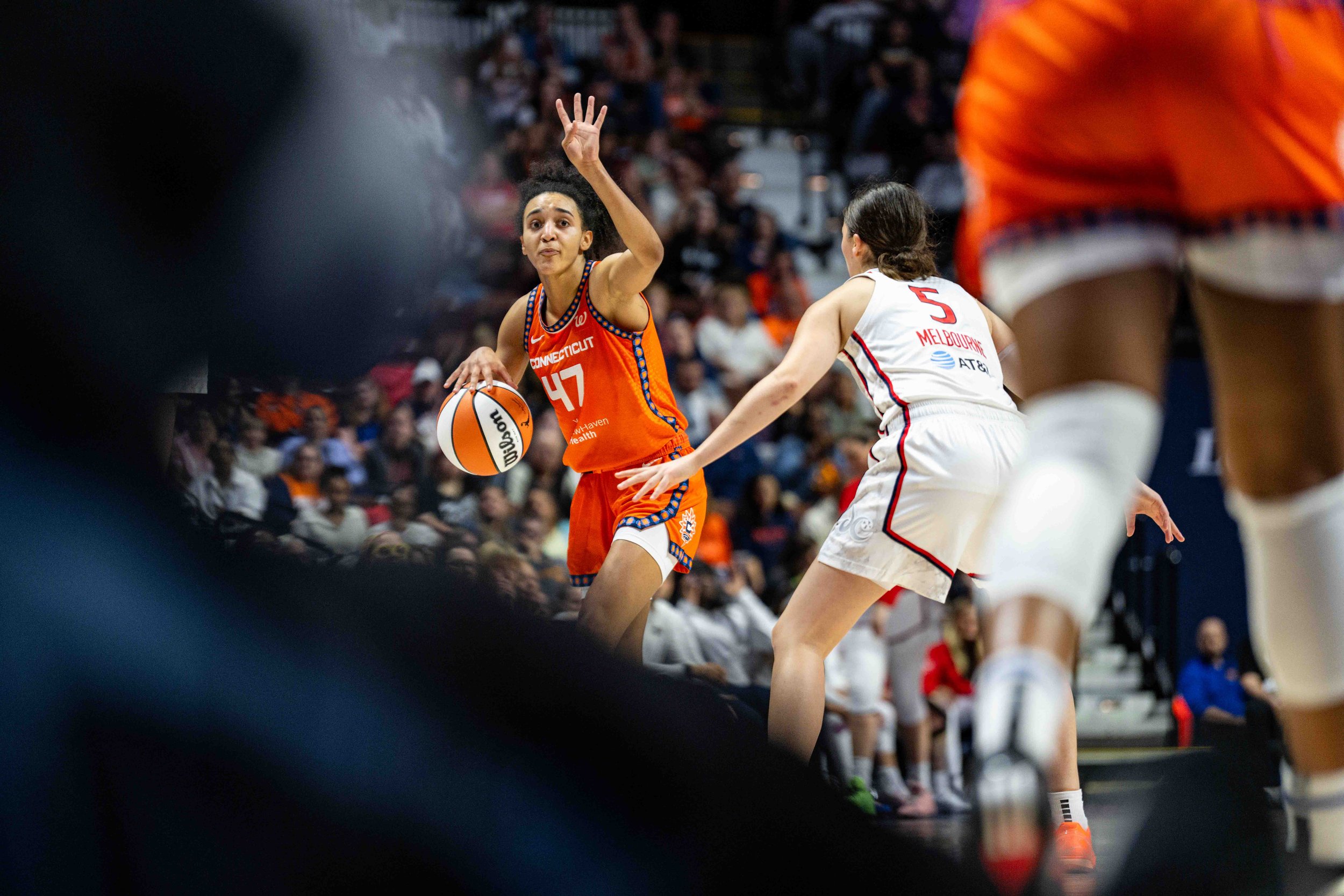 A women's basketball game with one player in a bright orange uniform holding a basketball and reaching upward, while a player in a white uniform with the number 5 defends. The background has a crowd of spectators.