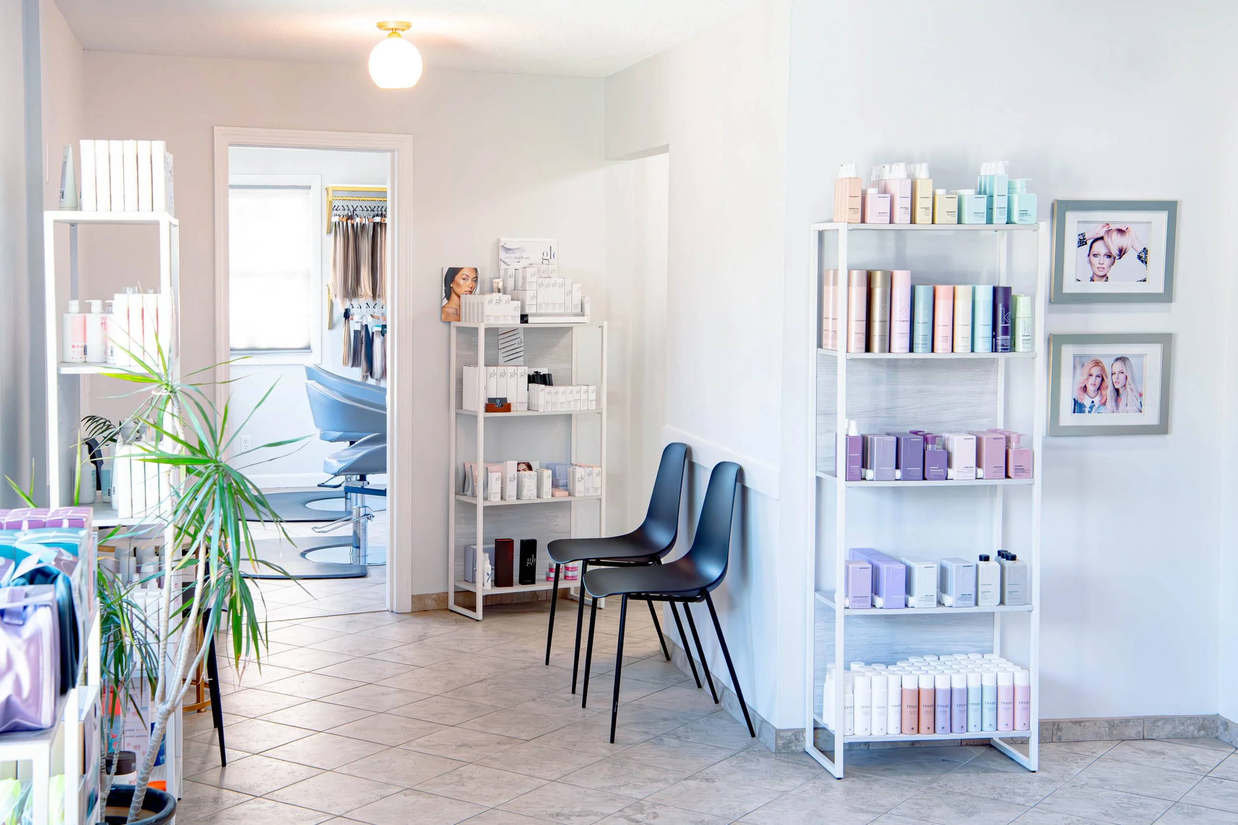 A bright, clean salon with two black chairs, white shelves filled with hair and beauty products, and framed pictures on the wall. In the background, a room with two salon chairs and a window.
