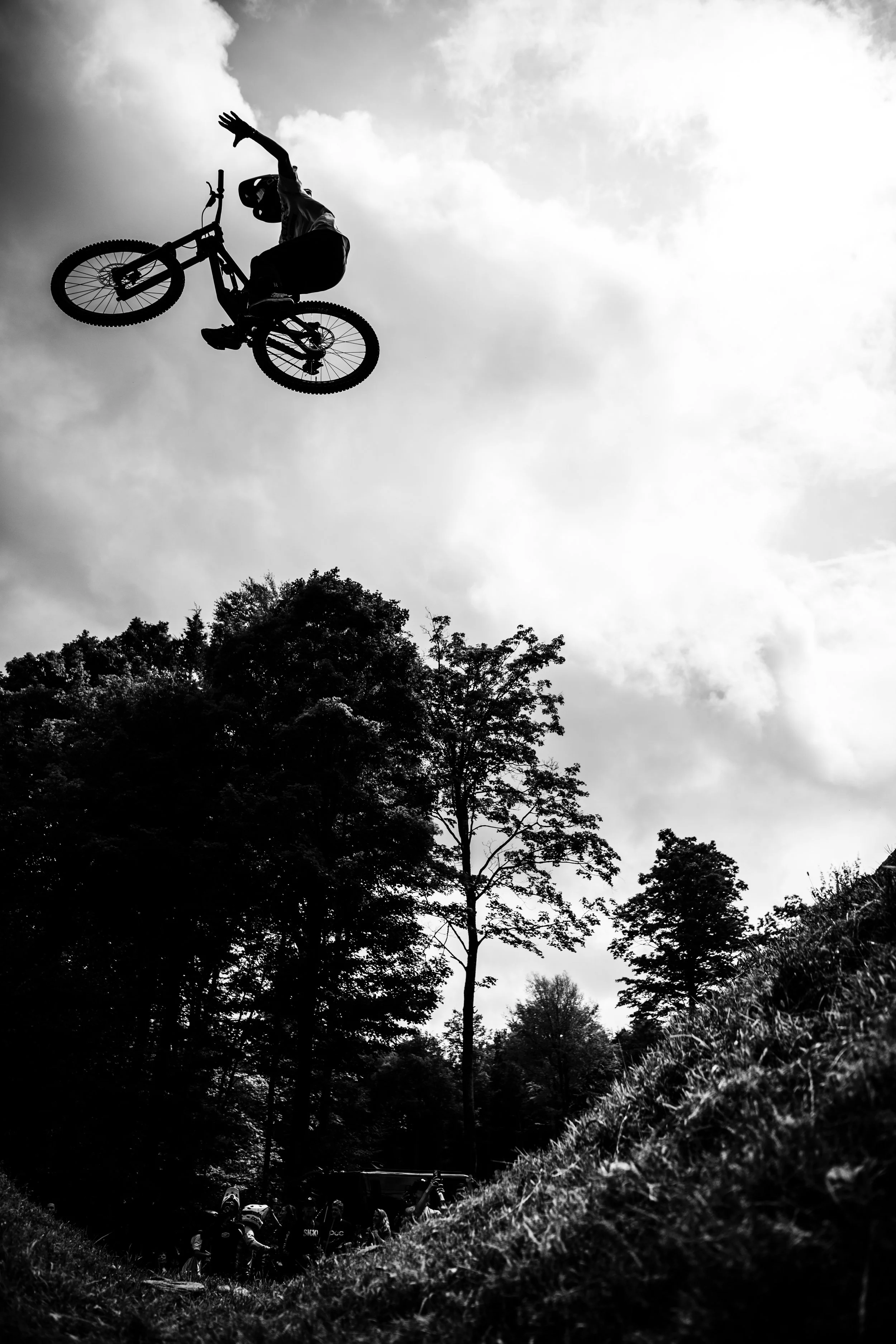 A person performing an aerial stunt on a mountain bike against a cloudy sky, with trees and a group of spectators below.