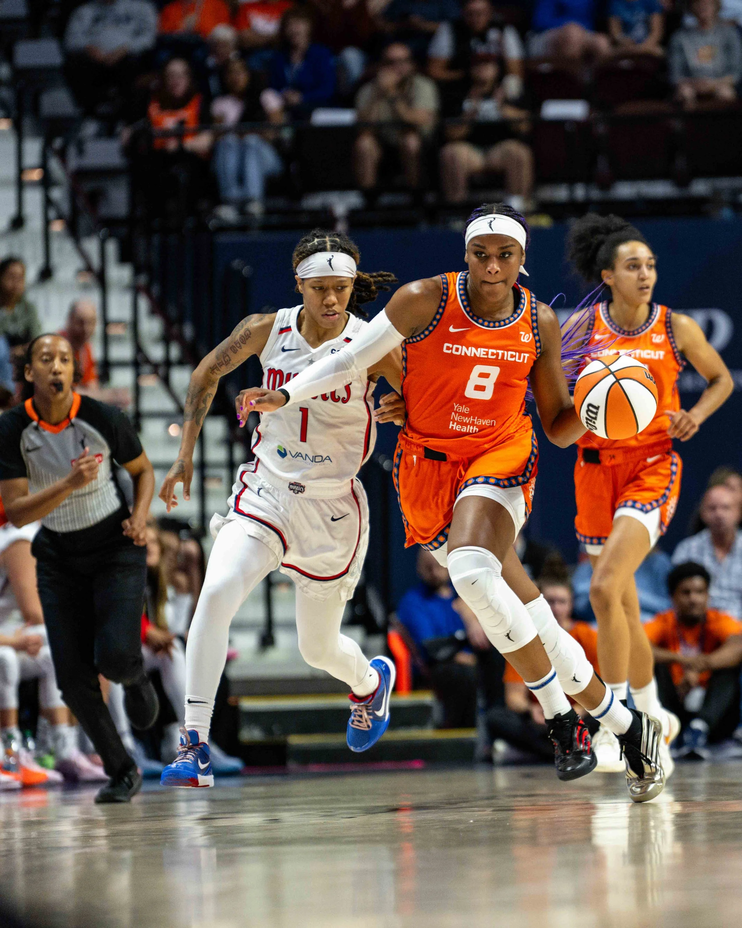 Women playing basketball in a game, with one player from Connecticut wearing an orange uniform dribbling the ball and a player from Miami in a white uniform defending. Referee and spectators in the background.