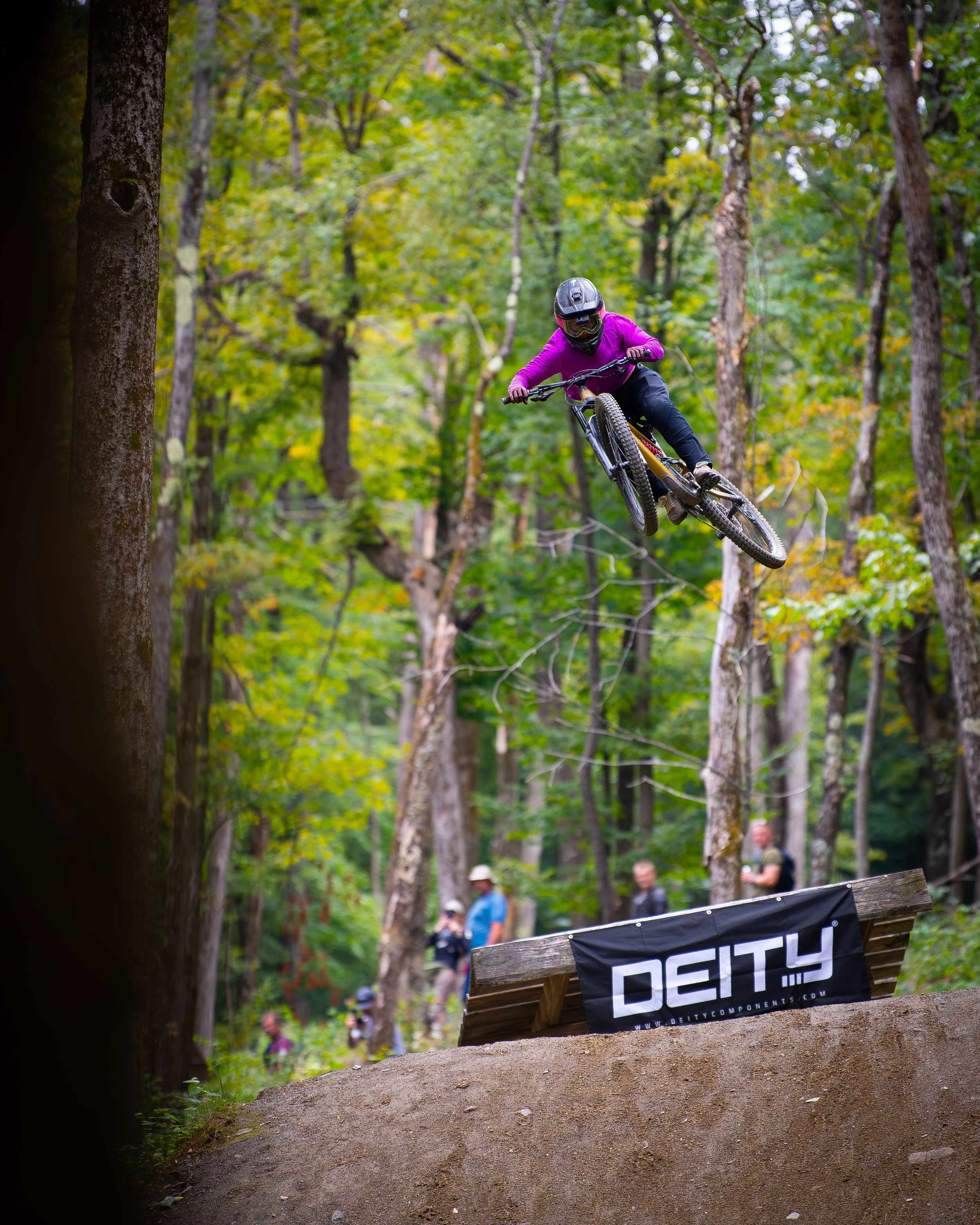 A mountain biker in a purple jacket, black helmet, and goggles is mid-air performing a jump on a dirt trail in a forest with green leaves. There are people in the background watching.