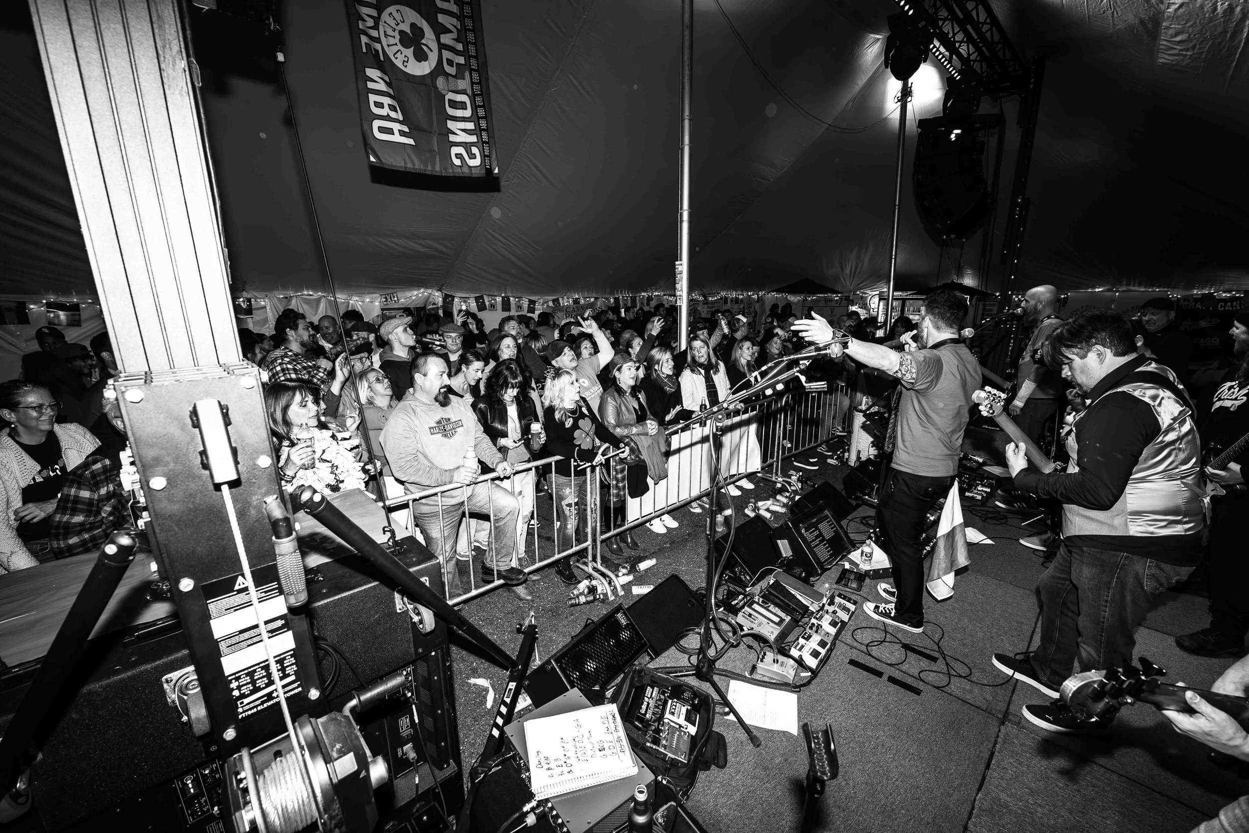 A black and white photo of a live music concert inside a large tent. The band is performing on stage with guitars, and the audience is standing behind a barrier, enjoying the show.