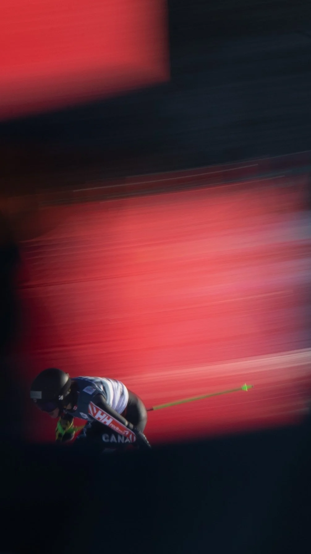 A skier in black gear and a white top, with a yellow pole, skiing downhill against a blurred red background.