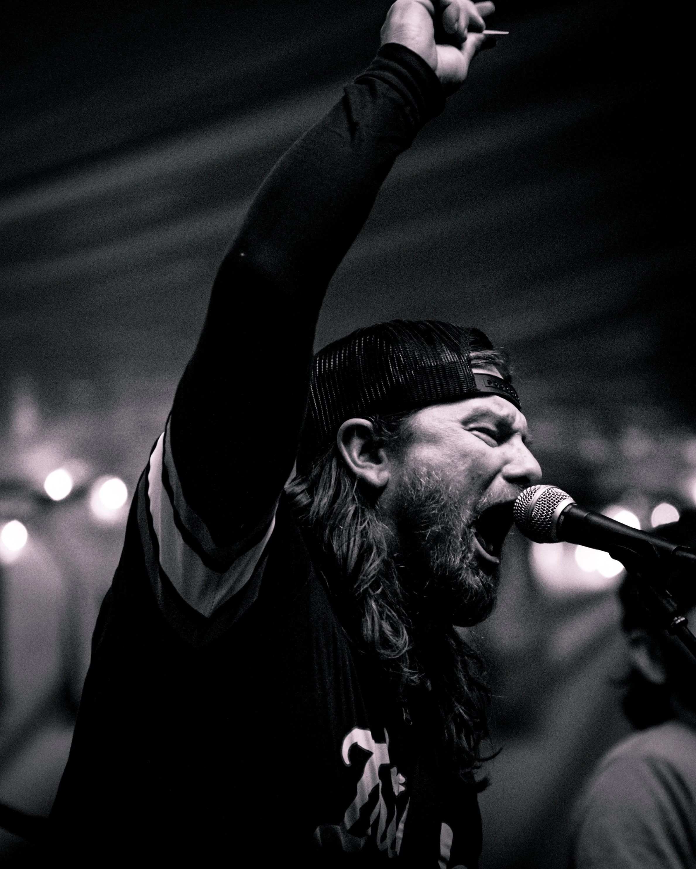 A man with long hair and a beard, wearing a baseball cap backwards, passionately singing into a microphone on stage, with his hand raised in the air.