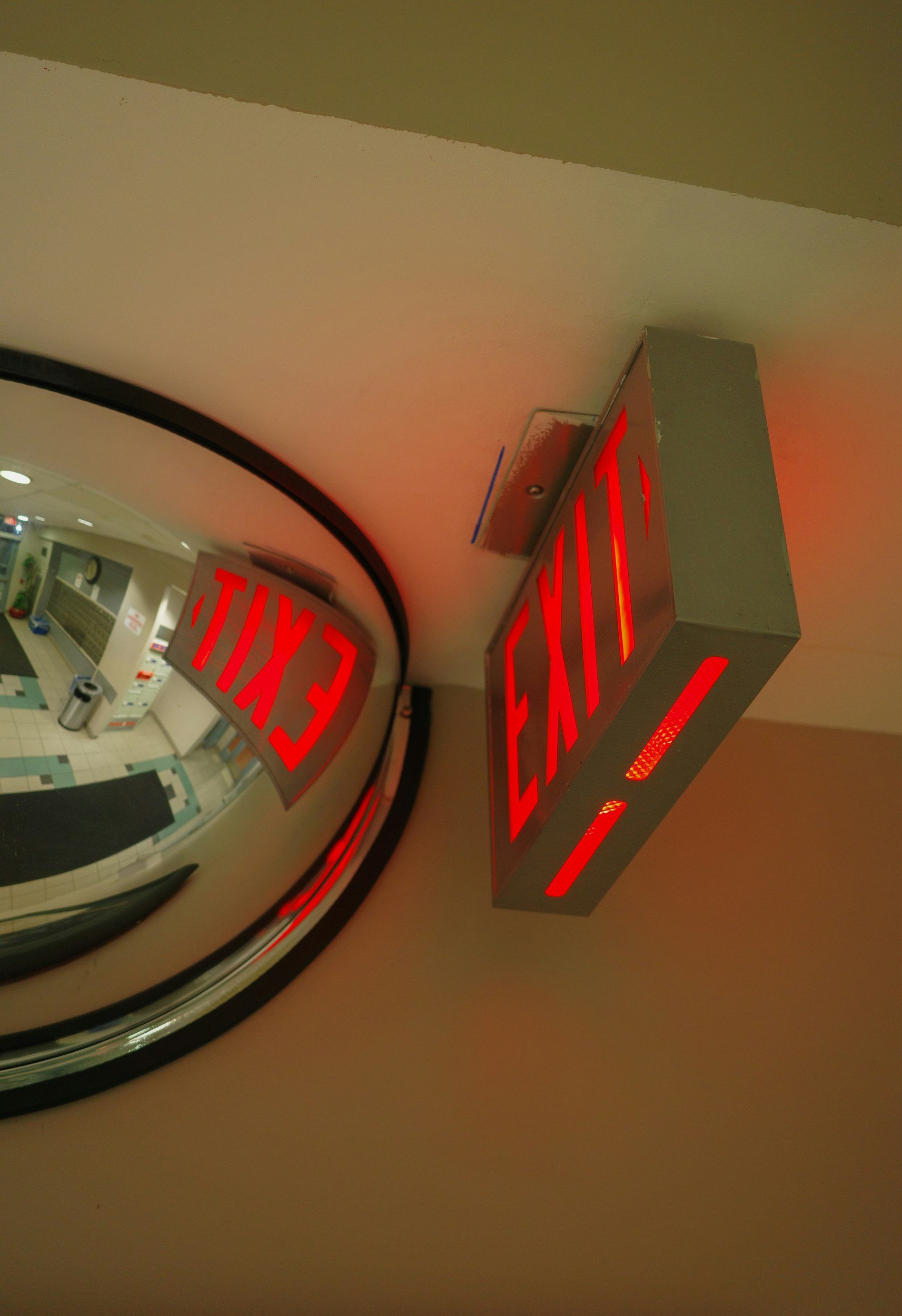 An illuminated red exit sign with the word 'EXIT' pointing to the right, mounted on a ceiling next to a reflective, curved surface with the word 'EXIT' also reflected.