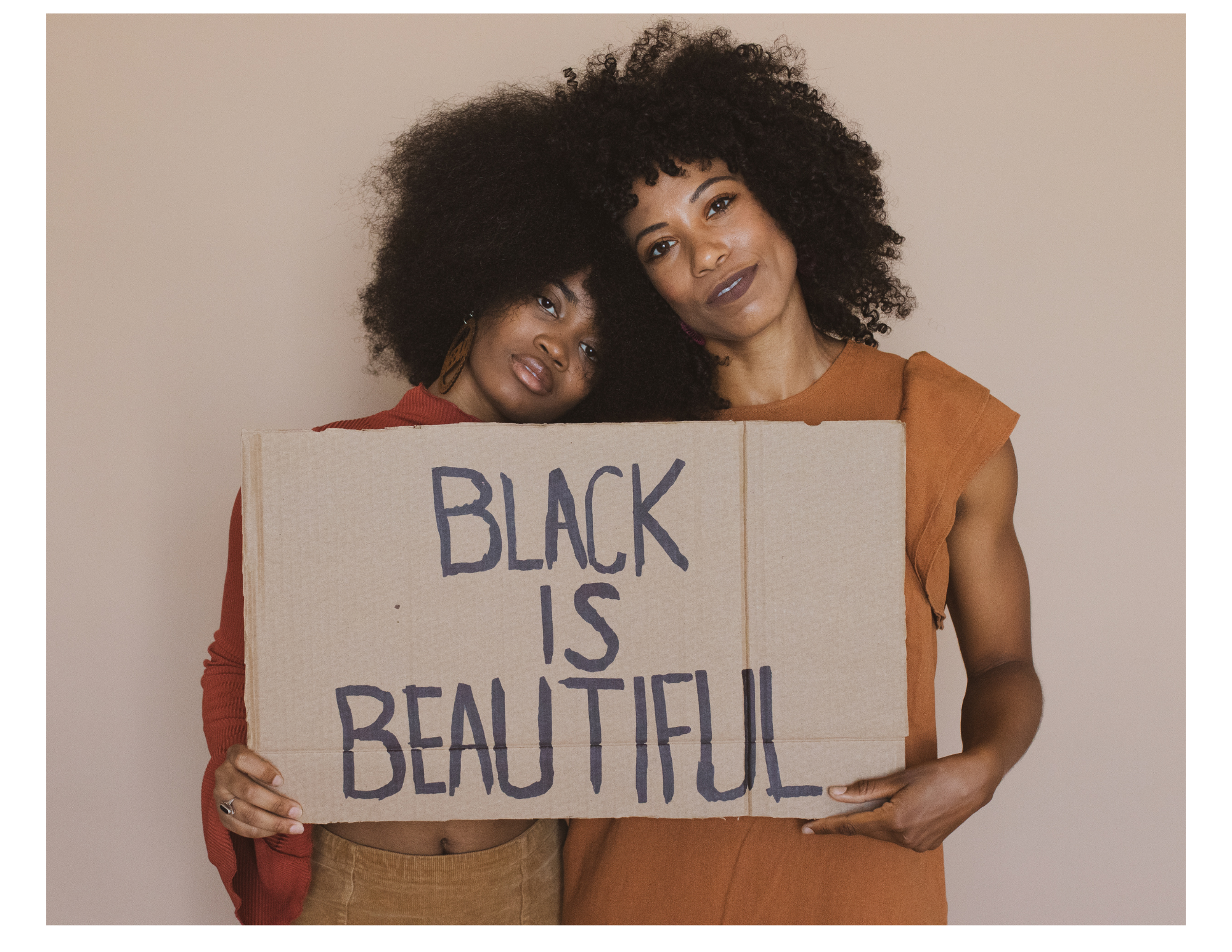 Two women with large curly hair, one with dark skin and one with lighter skin, stand closely together holding a handmade cardboard sign that reads 'Black is Beautiful.' They are smiling softly and leaning into each other, with a plain beige background.