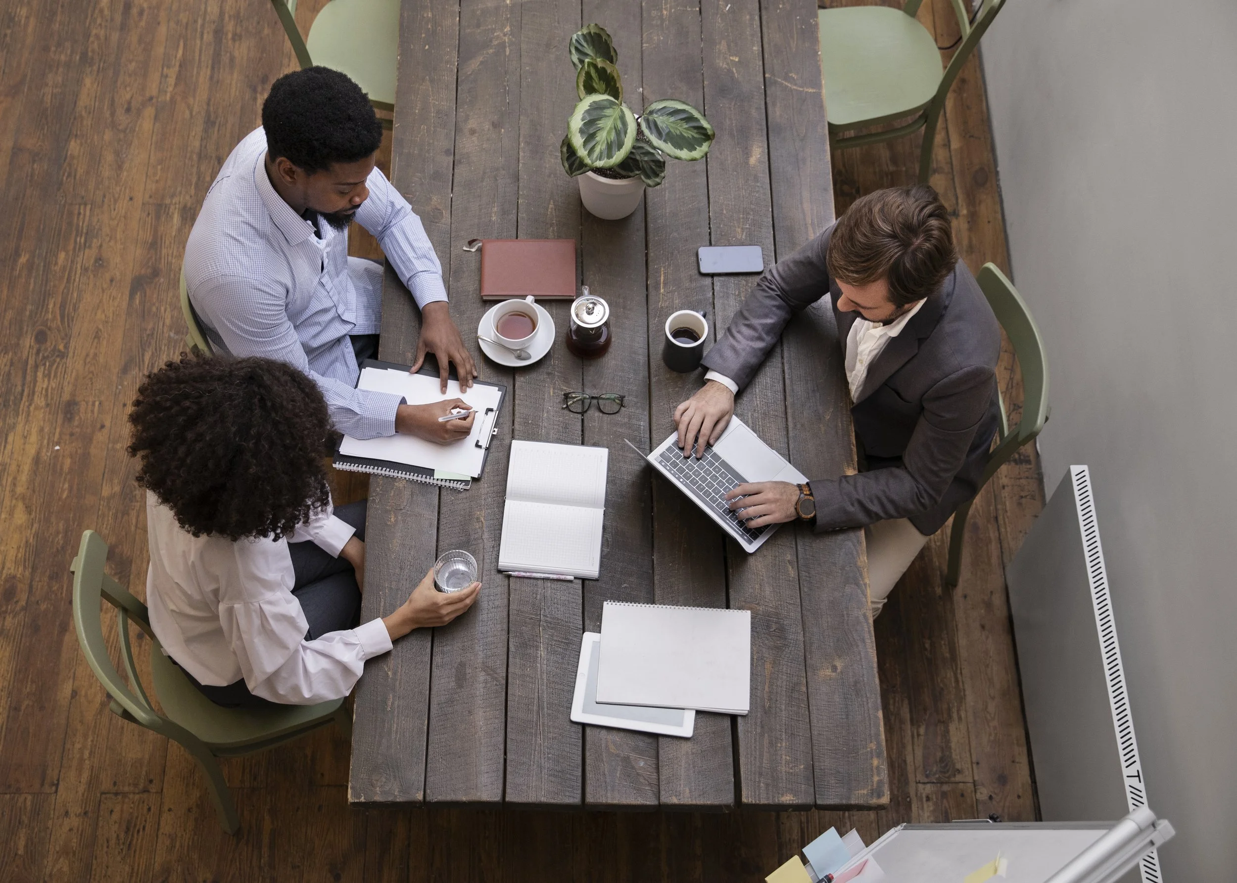 Two men and a woman sitting around a wooden conference table engaged in a meeting. The woman is holding a glass of water, one man is using a laptop, and the other man is writing on a notepad. The table has notebooks, a plant, cups of coffee, a water bottle, a smartphone, and glasses.