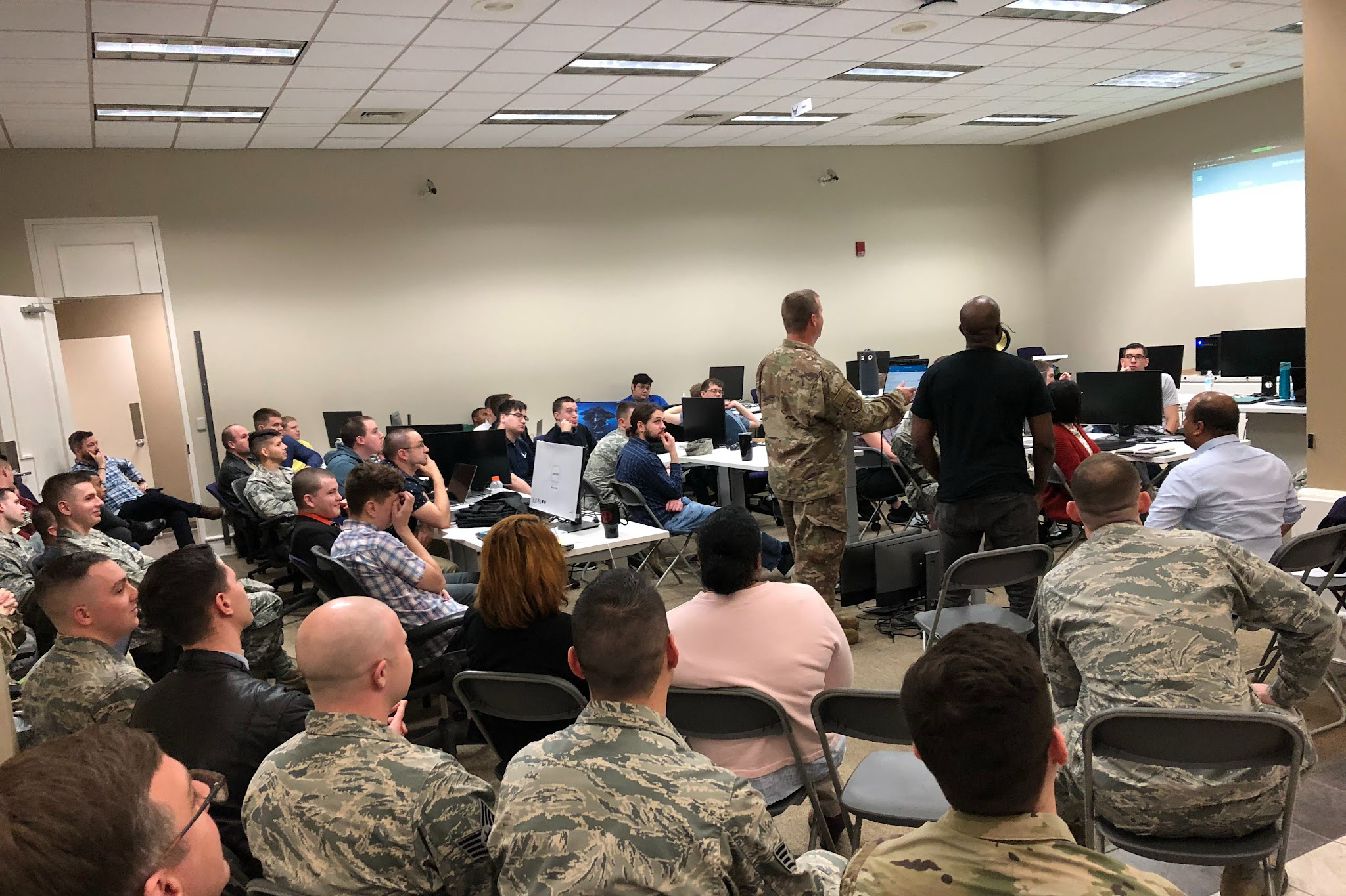 large meeting room with many people dressed in military and business casual clothing looking at a screen