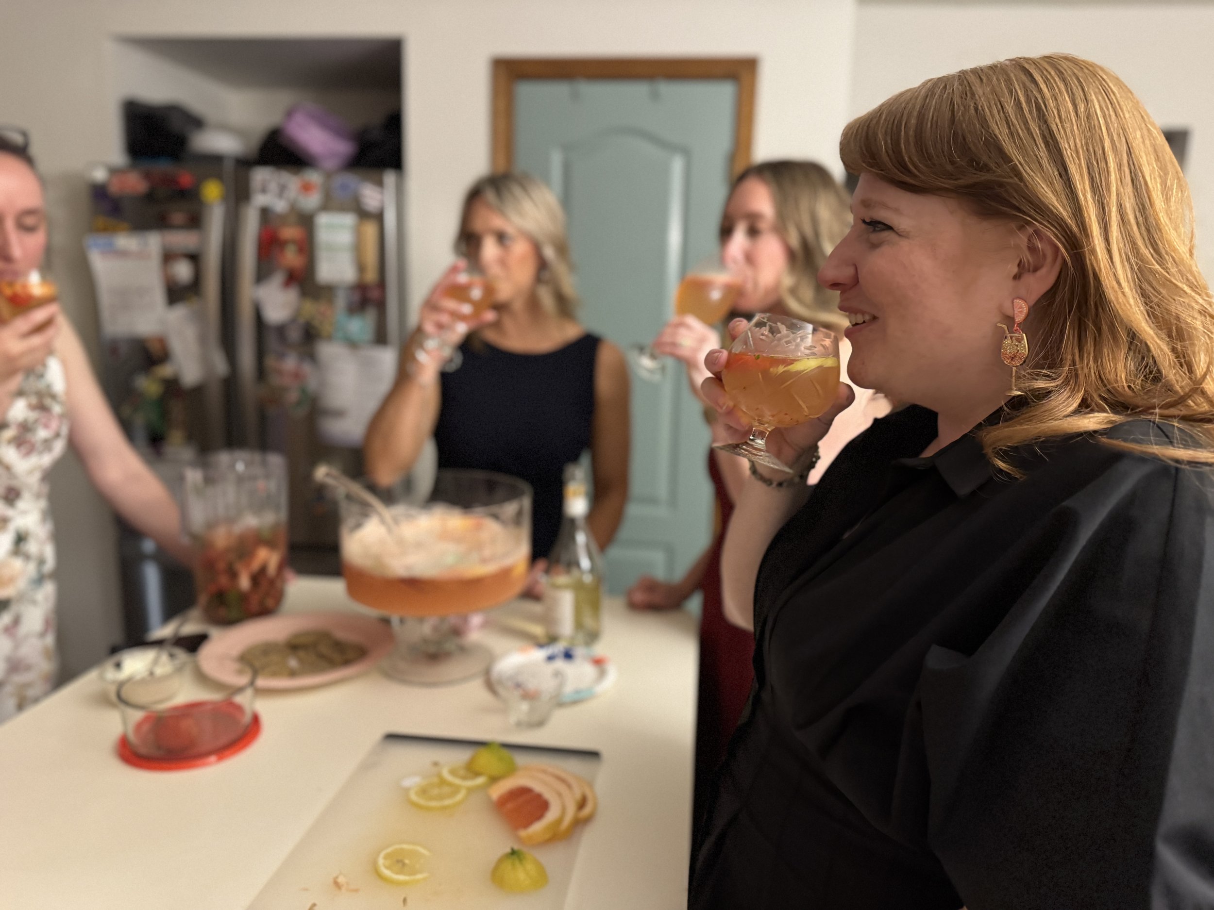 Four women at a gathering drinking cocktails in a home kitchen, with food and drinks on the table.