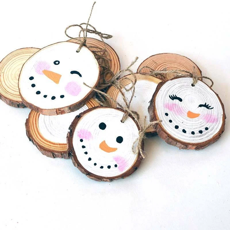 Decorative wood slices with snowman faces drawn on them, featuring eyes, noses, mouths, pink cheeks, and tied with twine bows.