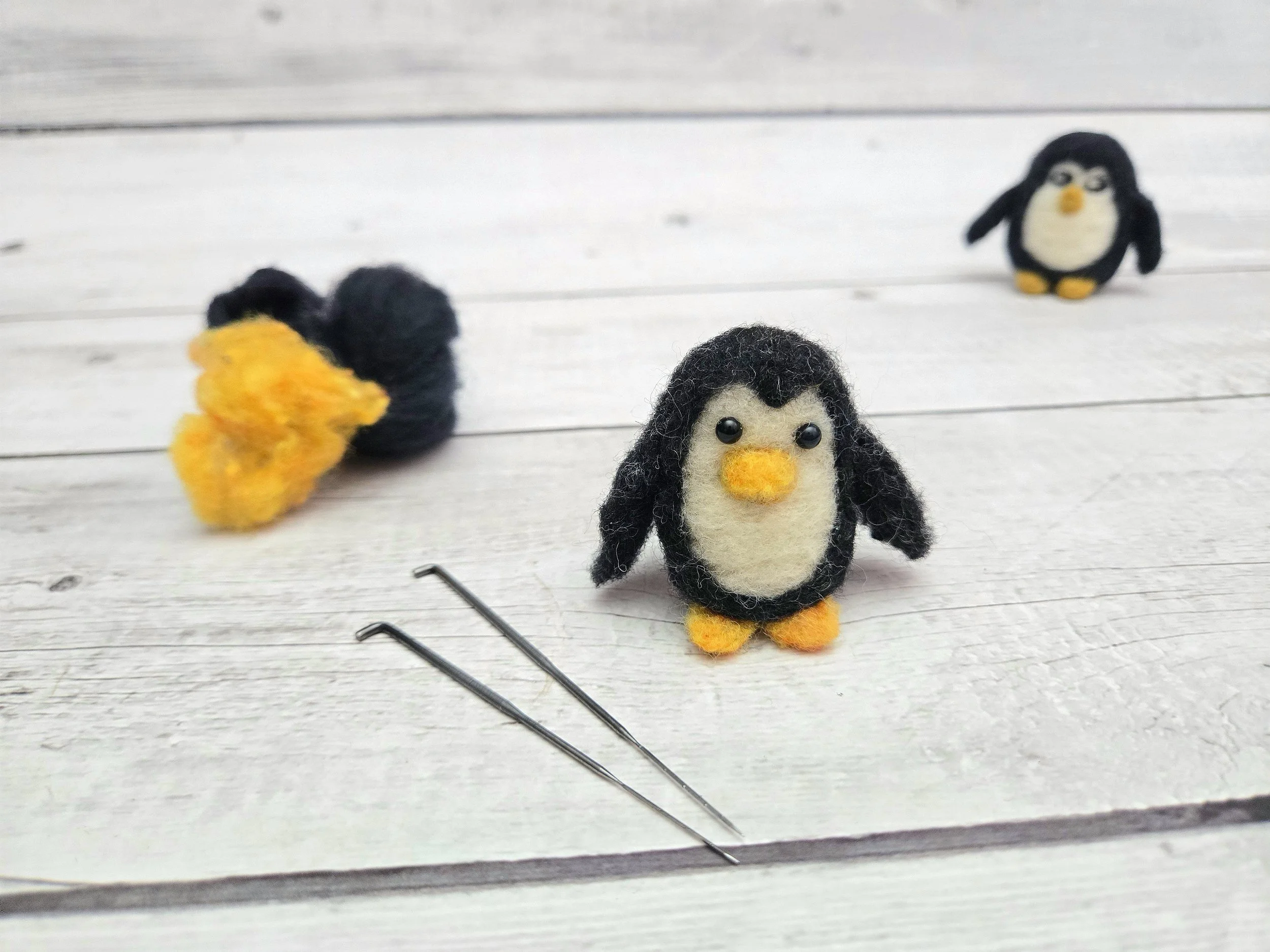 Three small felt penguin figures with orange beaks and feet on a white wooden surface, with two sewing pins lying nearby.