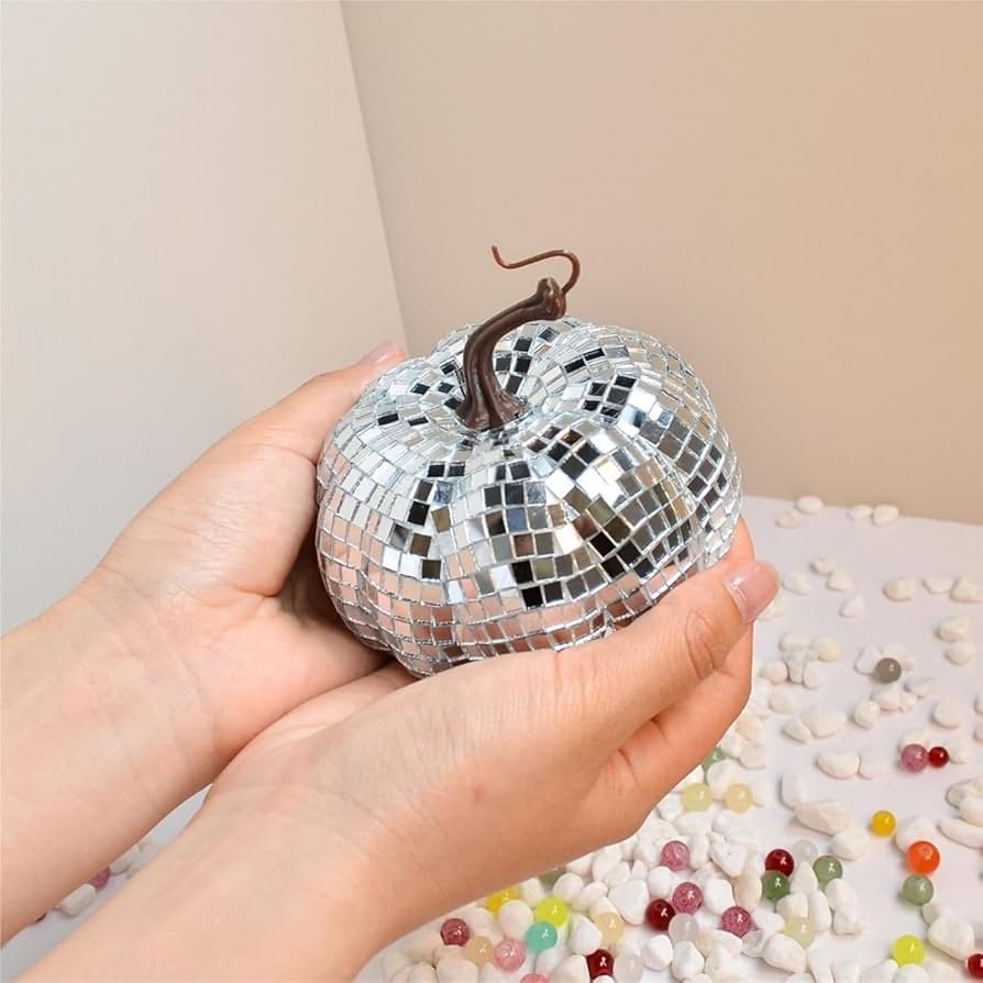 Person holding a small pumpkin decorated with silver mosaic tiles, with a stem, on a table covered with small colorful beads and white popcorn.