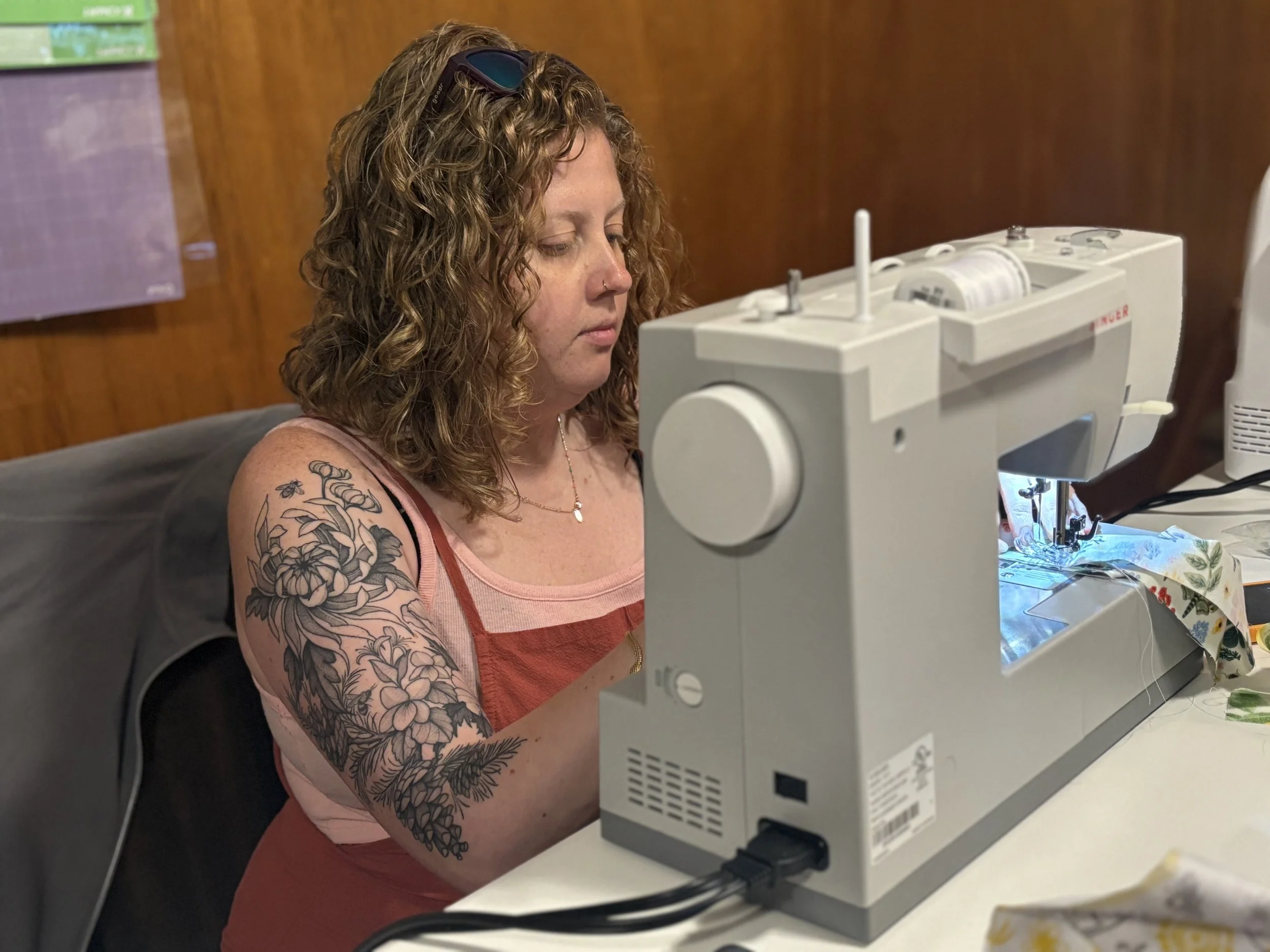 A woman with curly hair and a floral tattoo on her arm using a sewing machine.