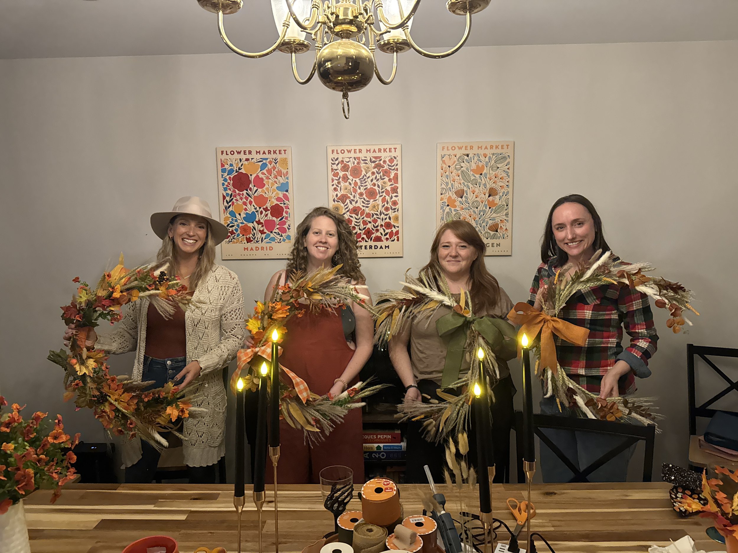 Four women standing behind a wooden table with fall-themed floral arrangements and decorative candles, smiling at the camera. The background features a white wall with three colorful poster prints labeled 'Flower Market'.