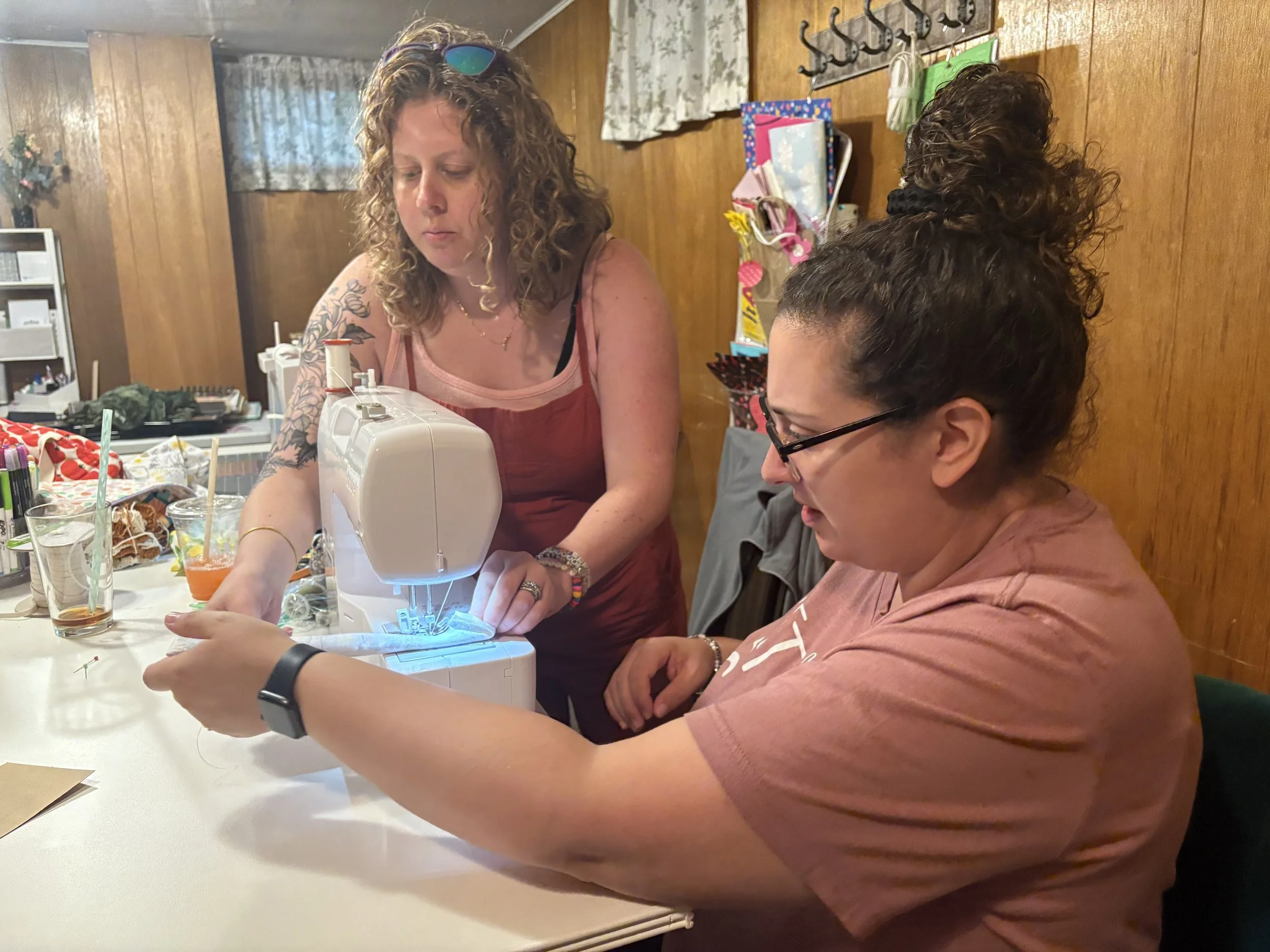 Two women working together on a sewing project at a table, one guiding the fabric through a sewing machine, with crafting supplies and drinks on the table in a cozy, wood-paneled room.