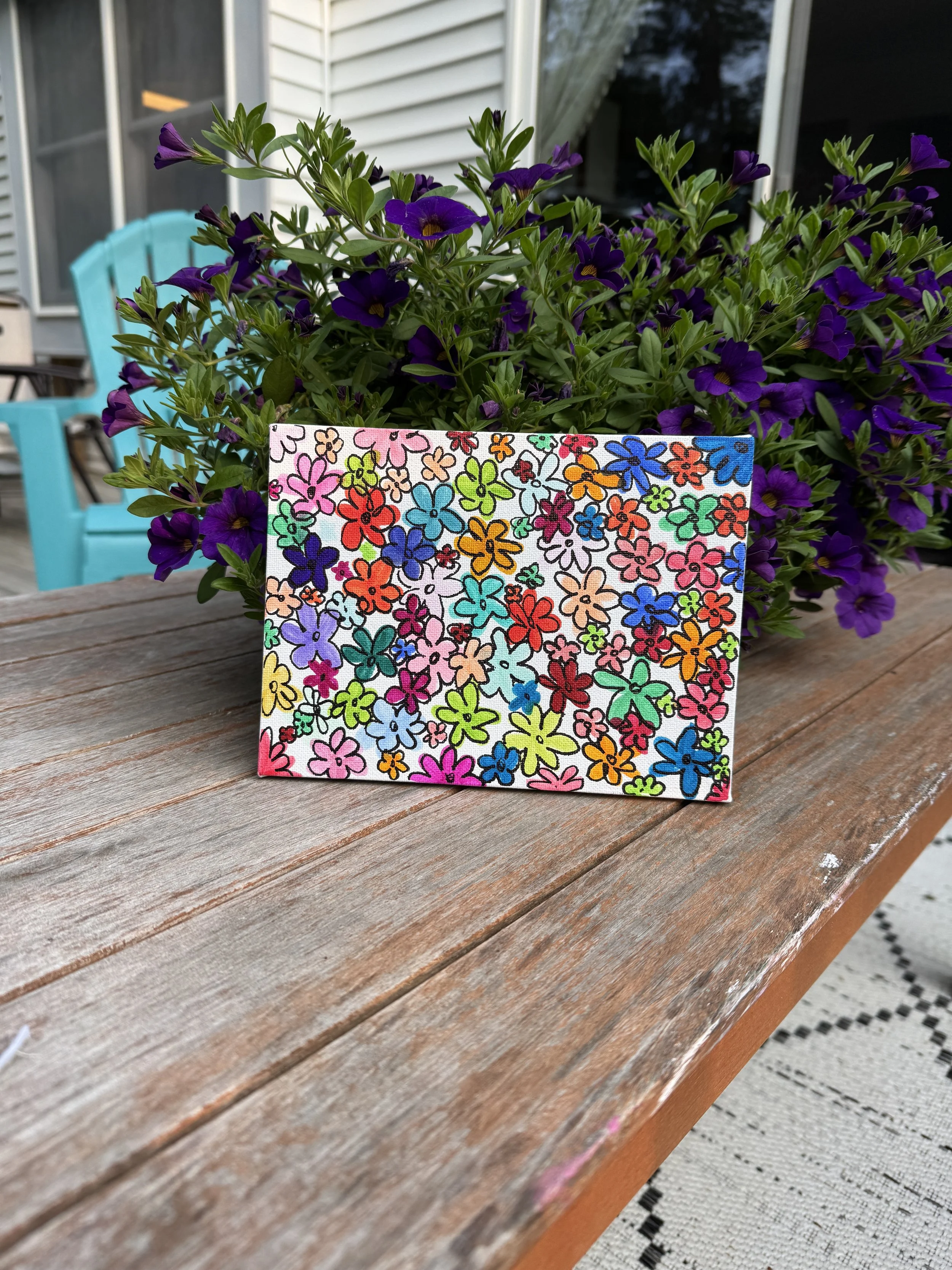 Colorful floral drawing of pink, purple, blue, red, orange, yellow, green, and peach flowers placed on a wooden table with purple petunias in the background.