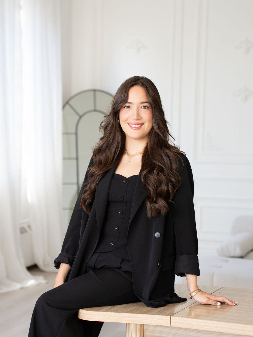 Mujer joven con cabello largo y rizado, vestida con ropa negra, sonriendo, en interiores con decoración blanca y elegante.