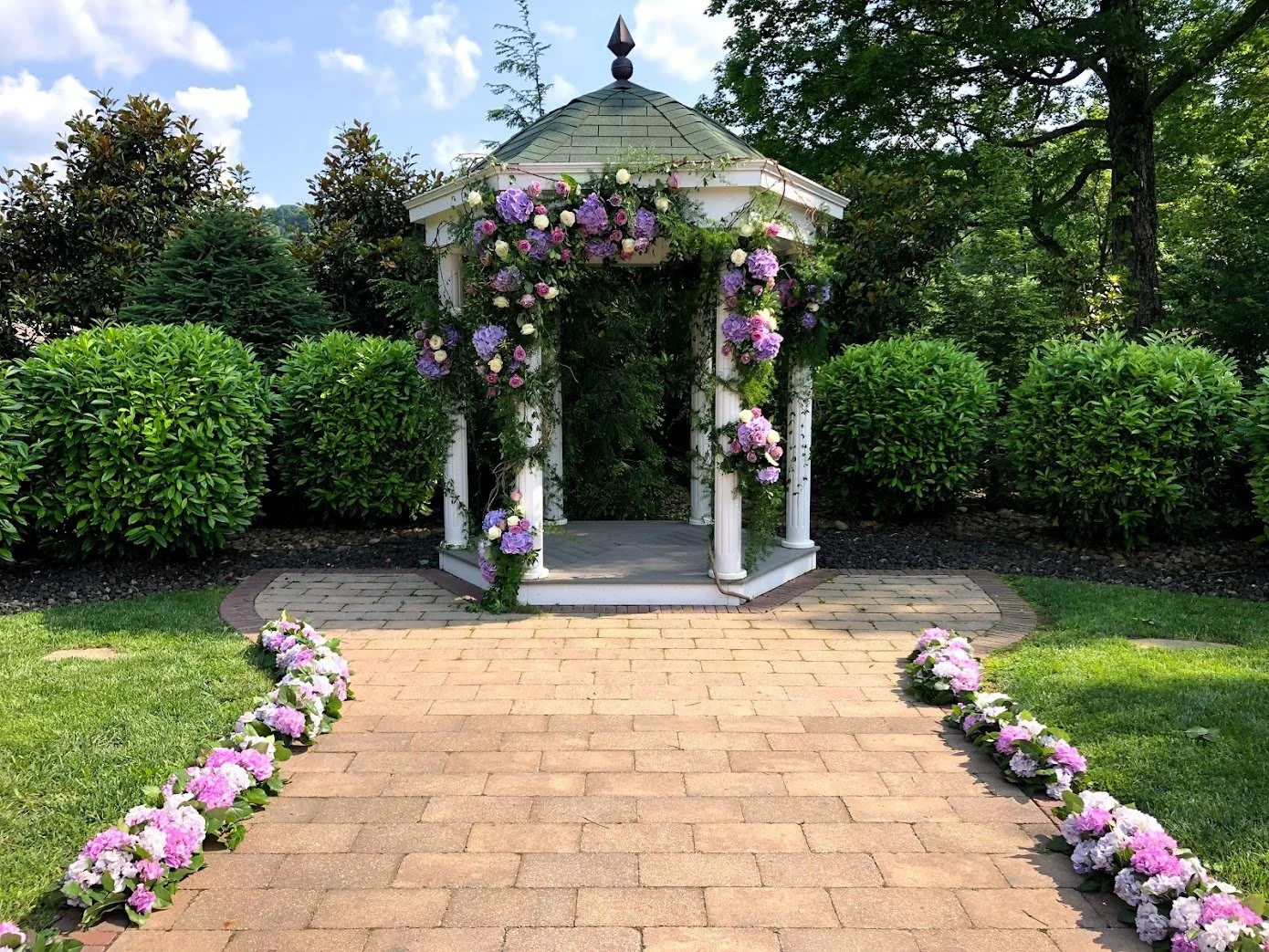 Good Services offering wedding and event florals in Des Moines Iowa showcasing a white gazebo decorated with purple and pink flowers, situated in a lush green garden with a brick pathway leading to it, surrounded by neatly trimmed bushes and trees.