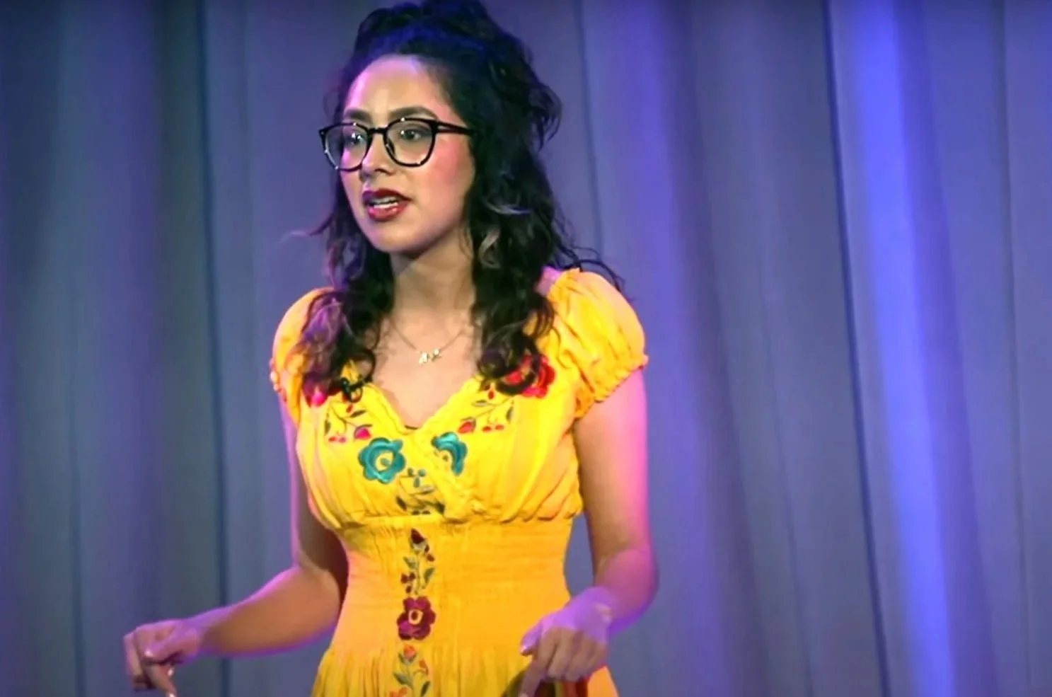A woman with dark curly hair, glasses, and wearing a yellow embroidered dress stands in front of a curtain backdrop.