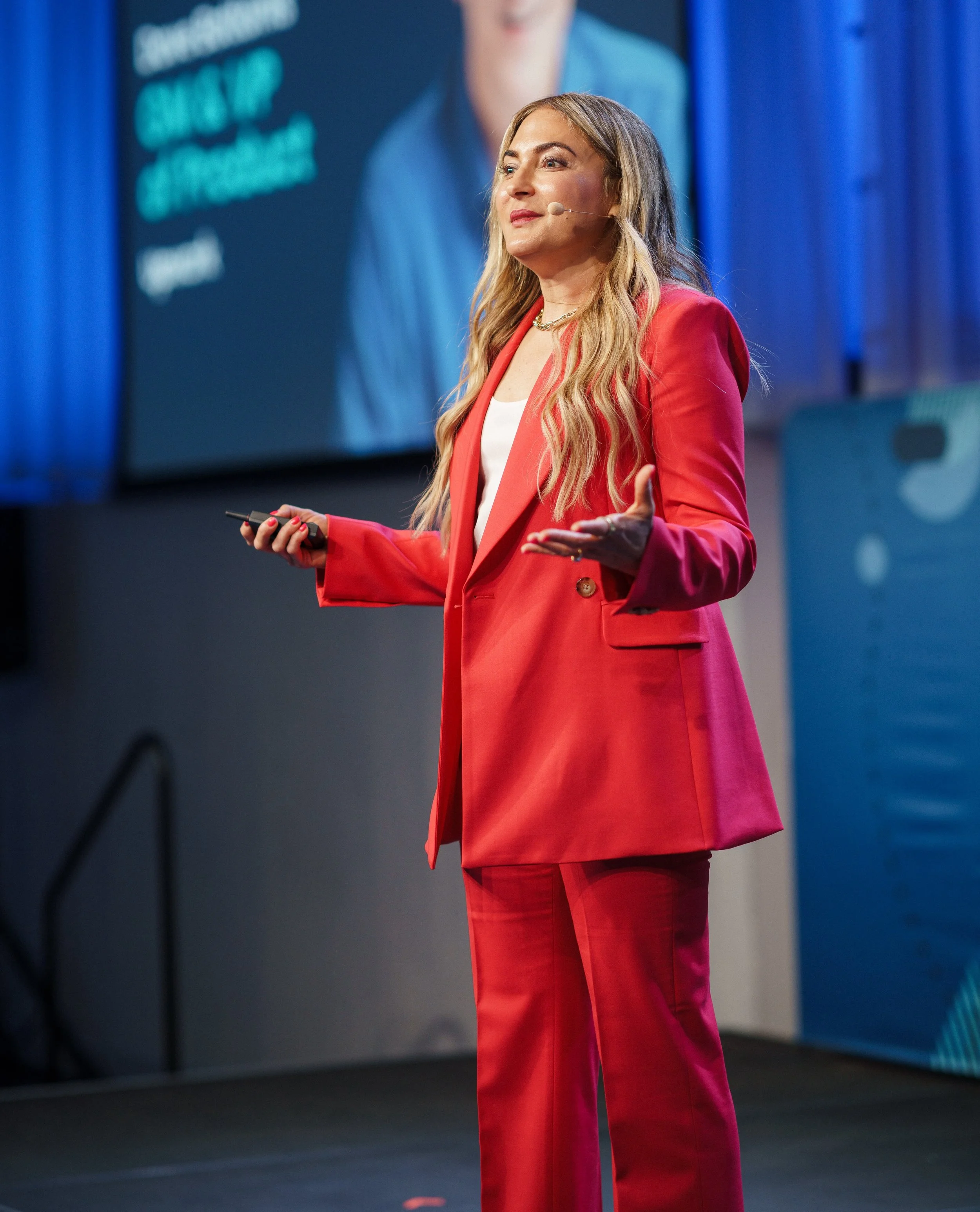 A woman in a red suit standing on stage giving a presentation, with a large screen behind her showing her image and some text.