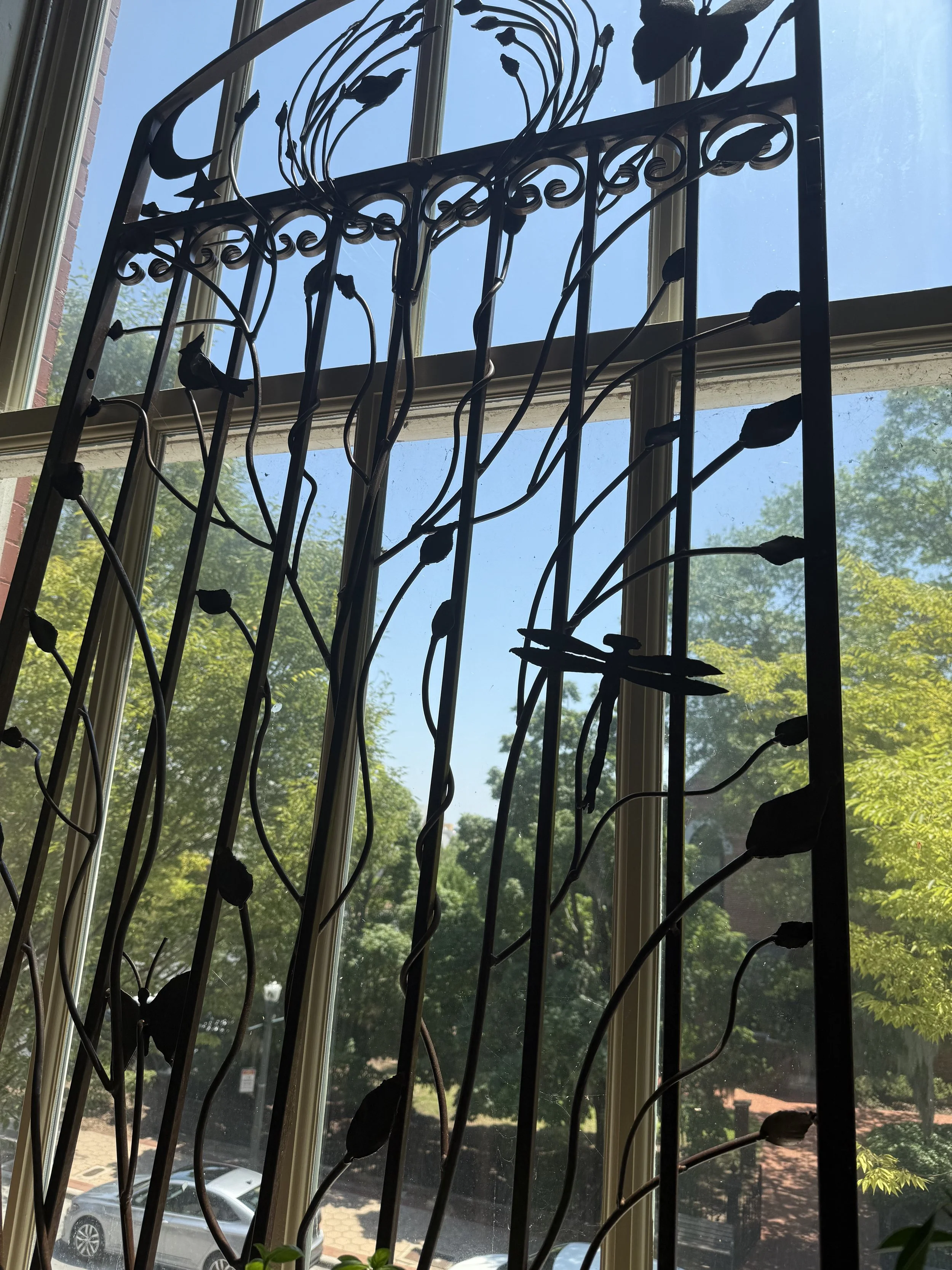 A decorative metal window grille with floral and leaf patterns, seen against a bright blue sky and green trees outdoors.