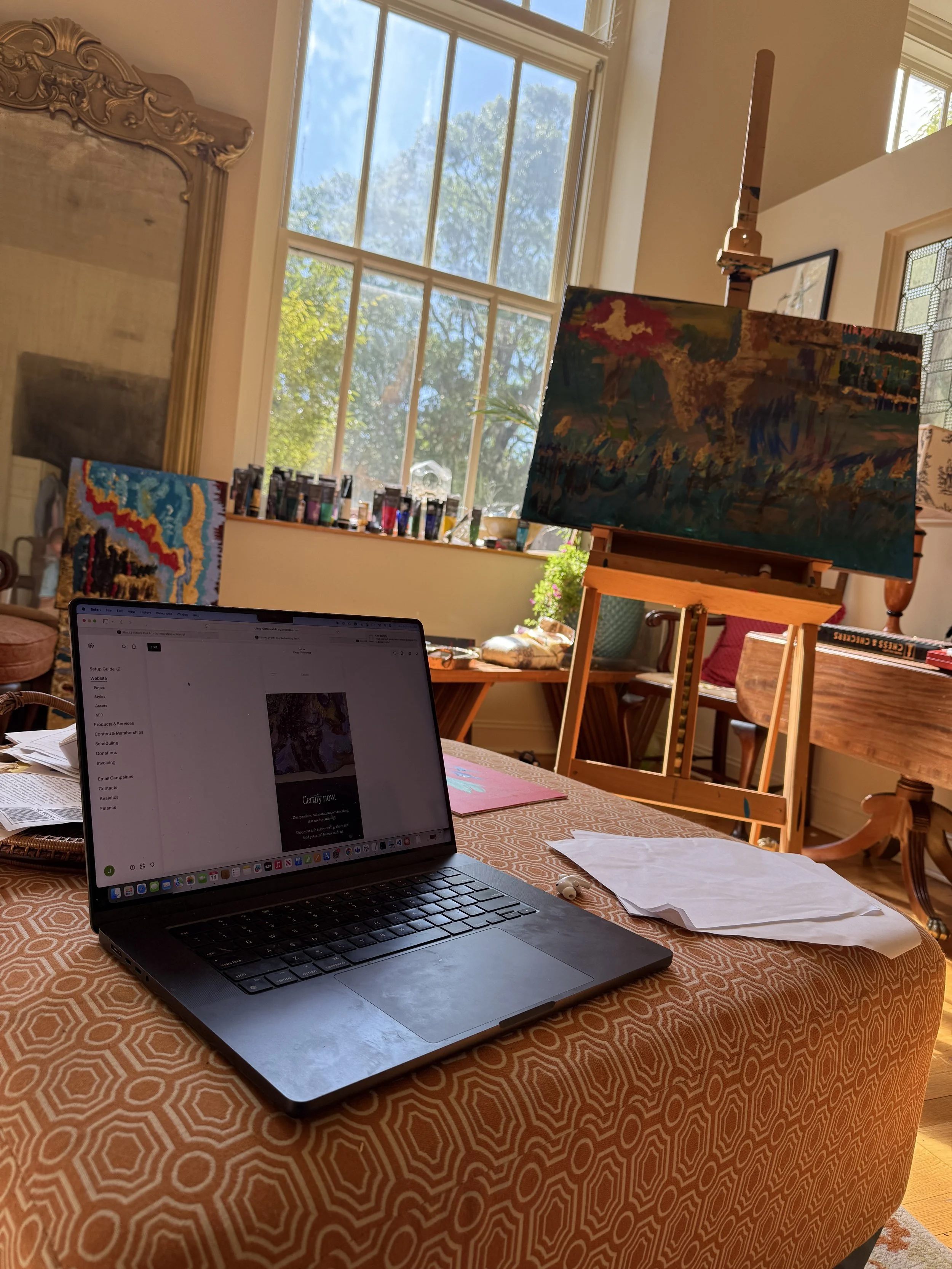 An art studio with a laptop on a patterned orange tablecloth, papers, and a paint palette. In the background, two colorful paintings are displayed, one on an easel, with a large window letting in natural light and trees outside.